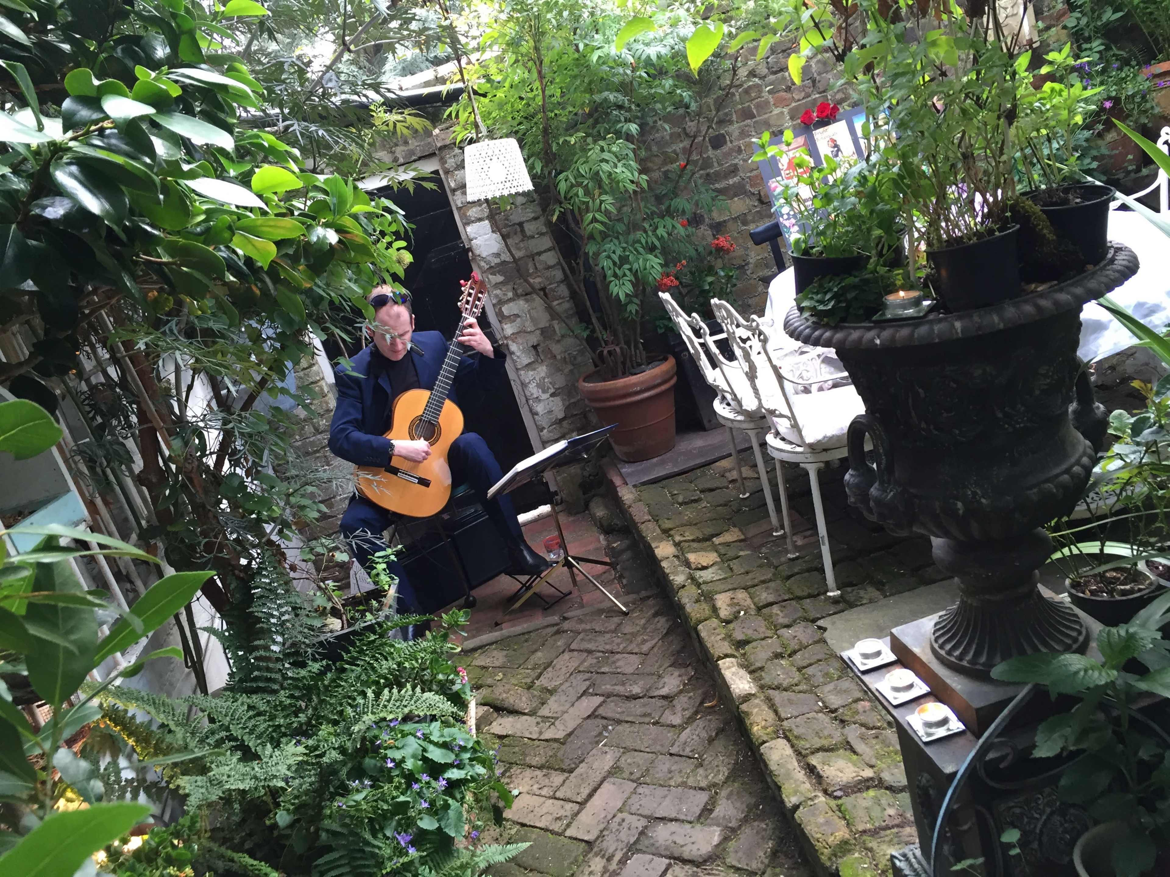 A musician plays a classical guitar in a lush garden courtyard filled with greenery and outdoor seating.