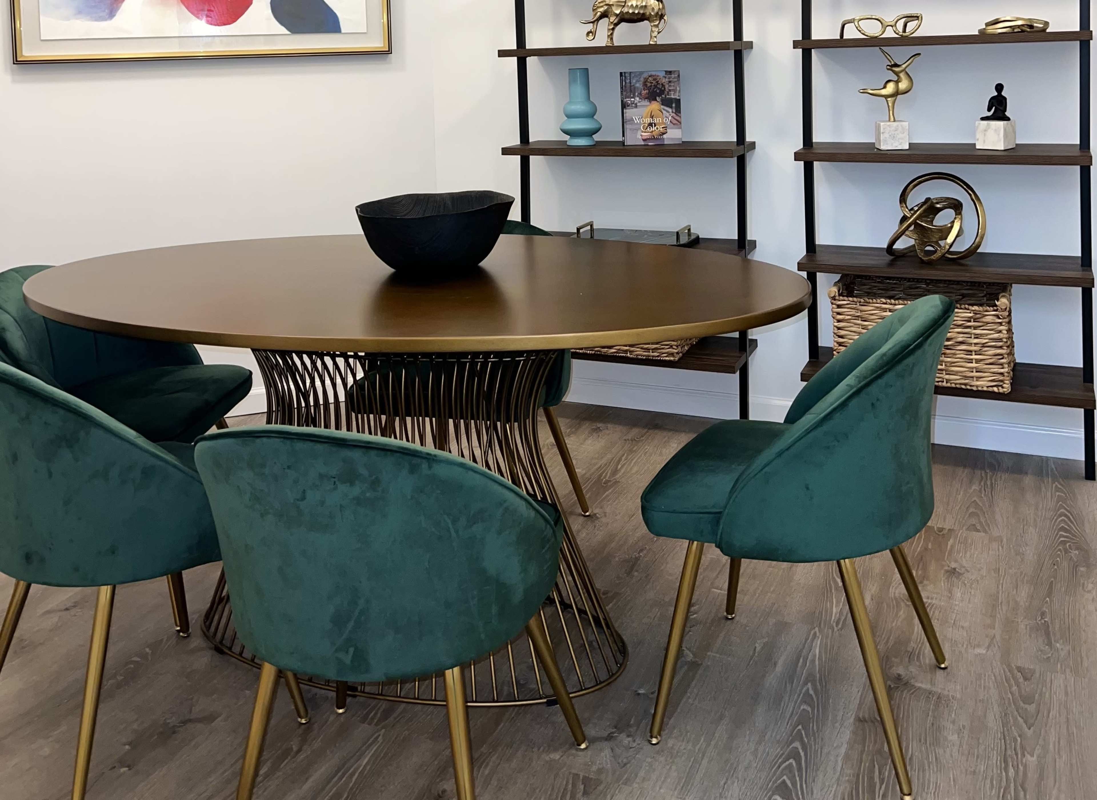 A round table with a metal base is surrounded by four green velvet chairs in a well-decorated room featuring shelves with various decorative items.
