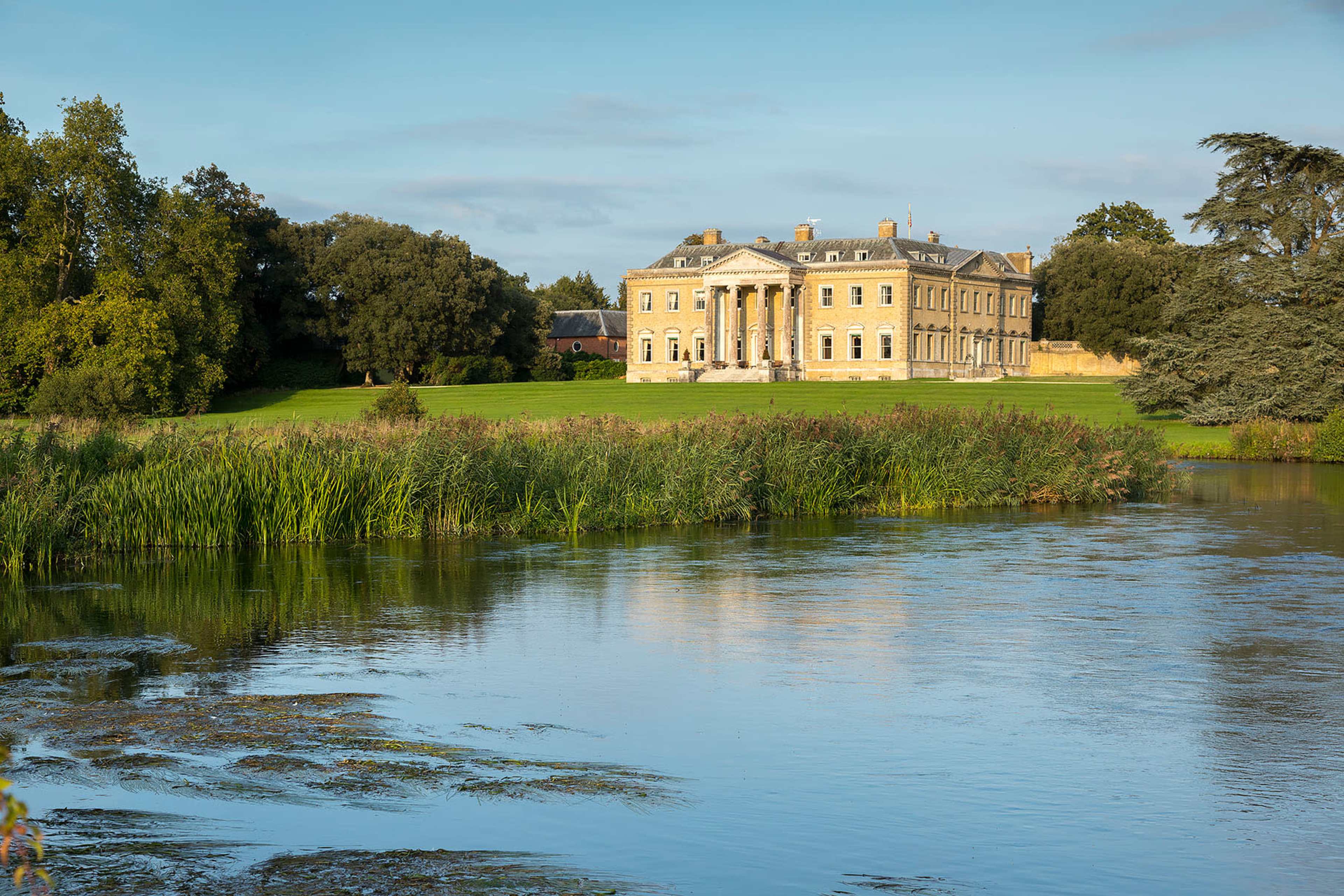 A large, classical-style mansion stands on a hillside near a calm river, surrounded by trees and manicured lawns.