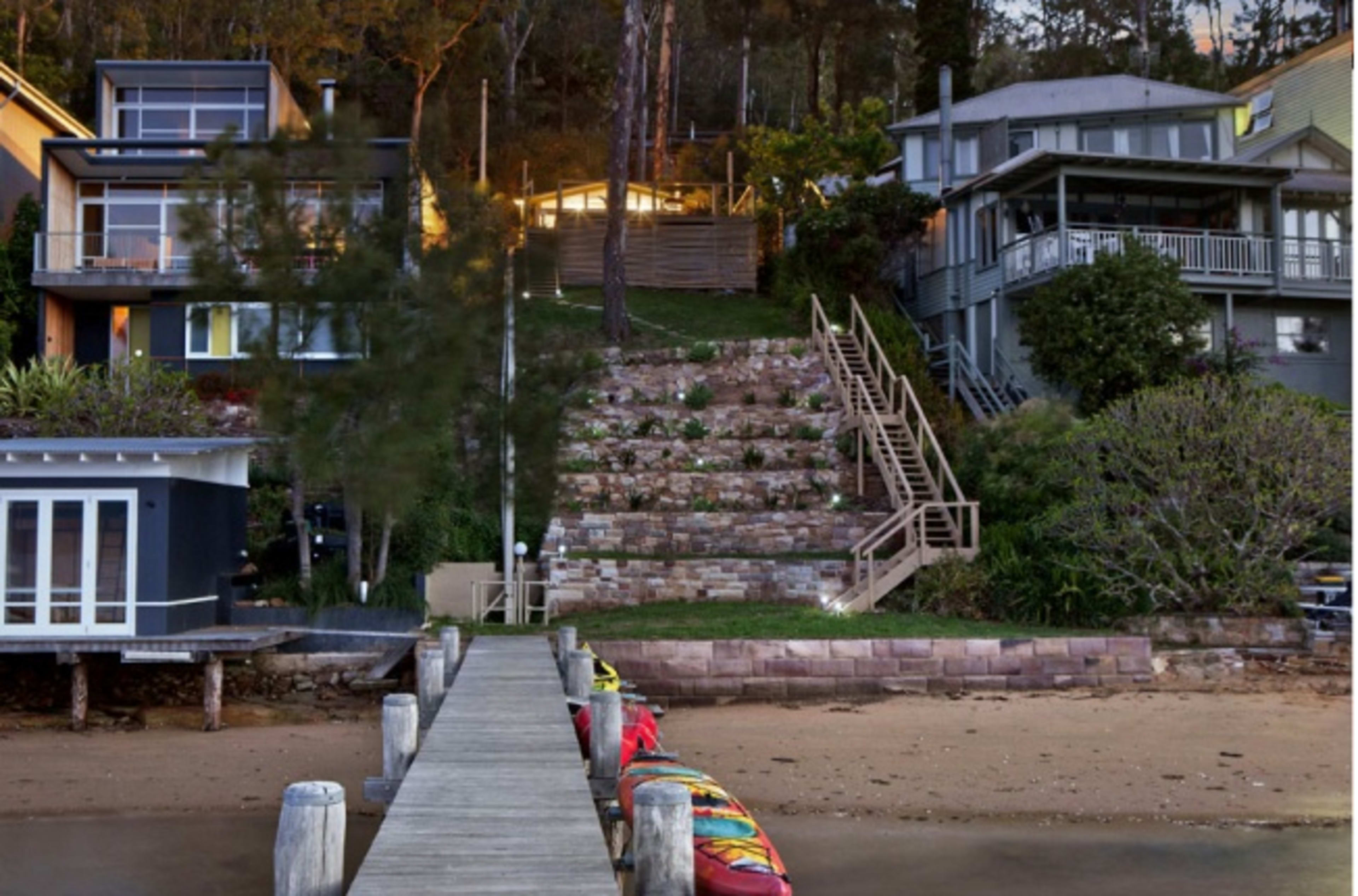 A waterfront view of a dock leading to a staircase that ascends to two houses, with lush greenery surrounding the area.
