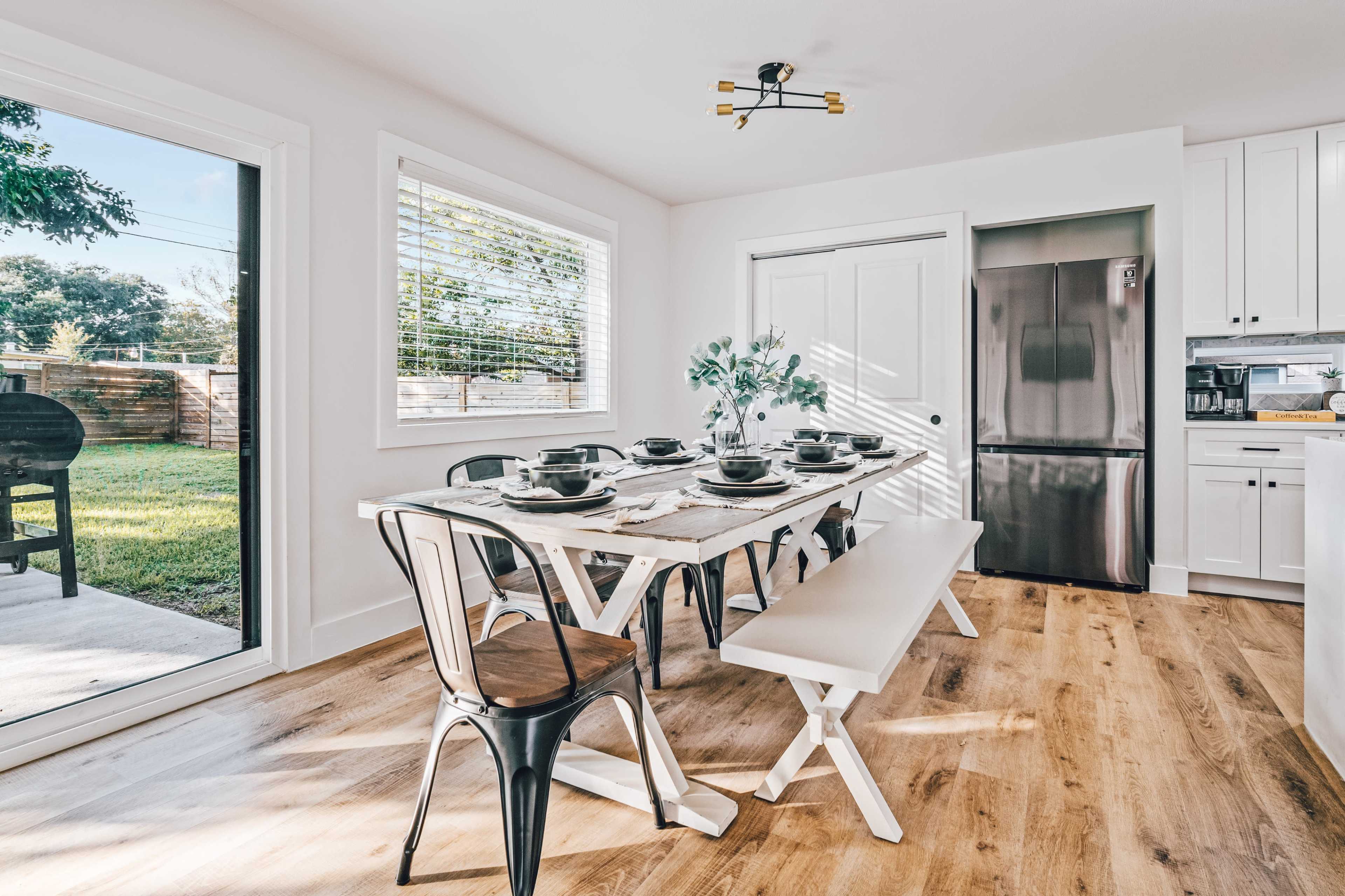 A dining area with a long table set for a meal, featuring black chairs, a wooden bench, and a view of a backyard through a sliding glass door.