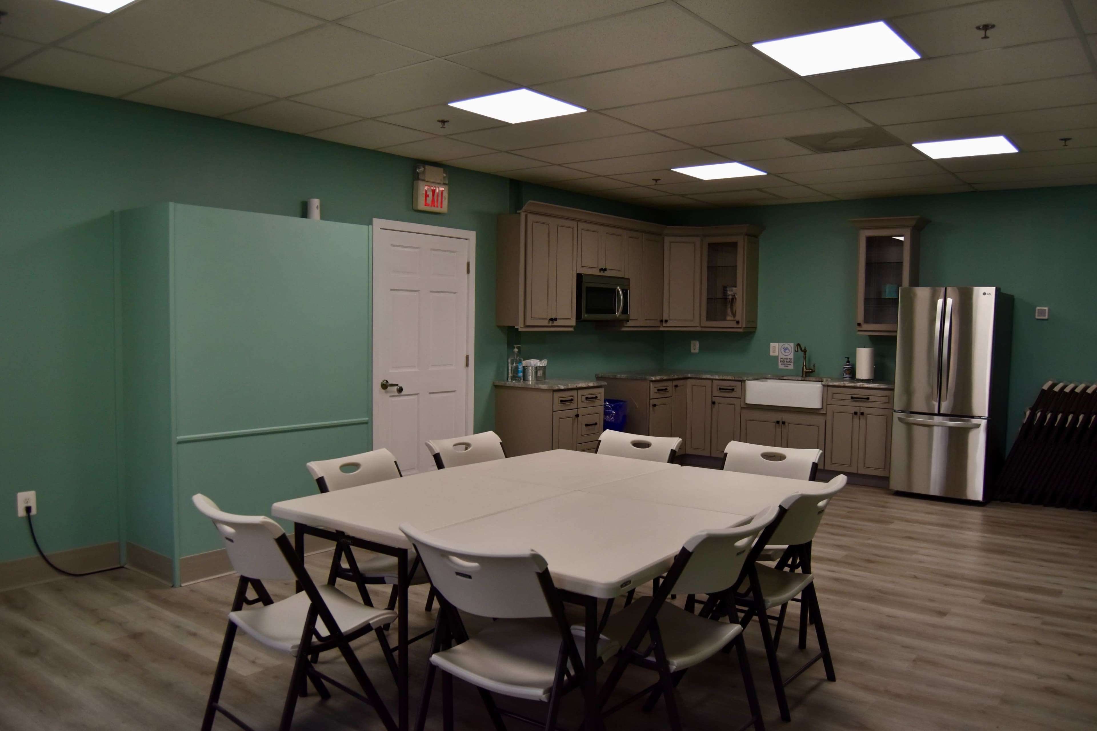 A well-lit kitchen area features a large rectangular table surrounded by folding chairs, with cabinets and appliances along the walls.