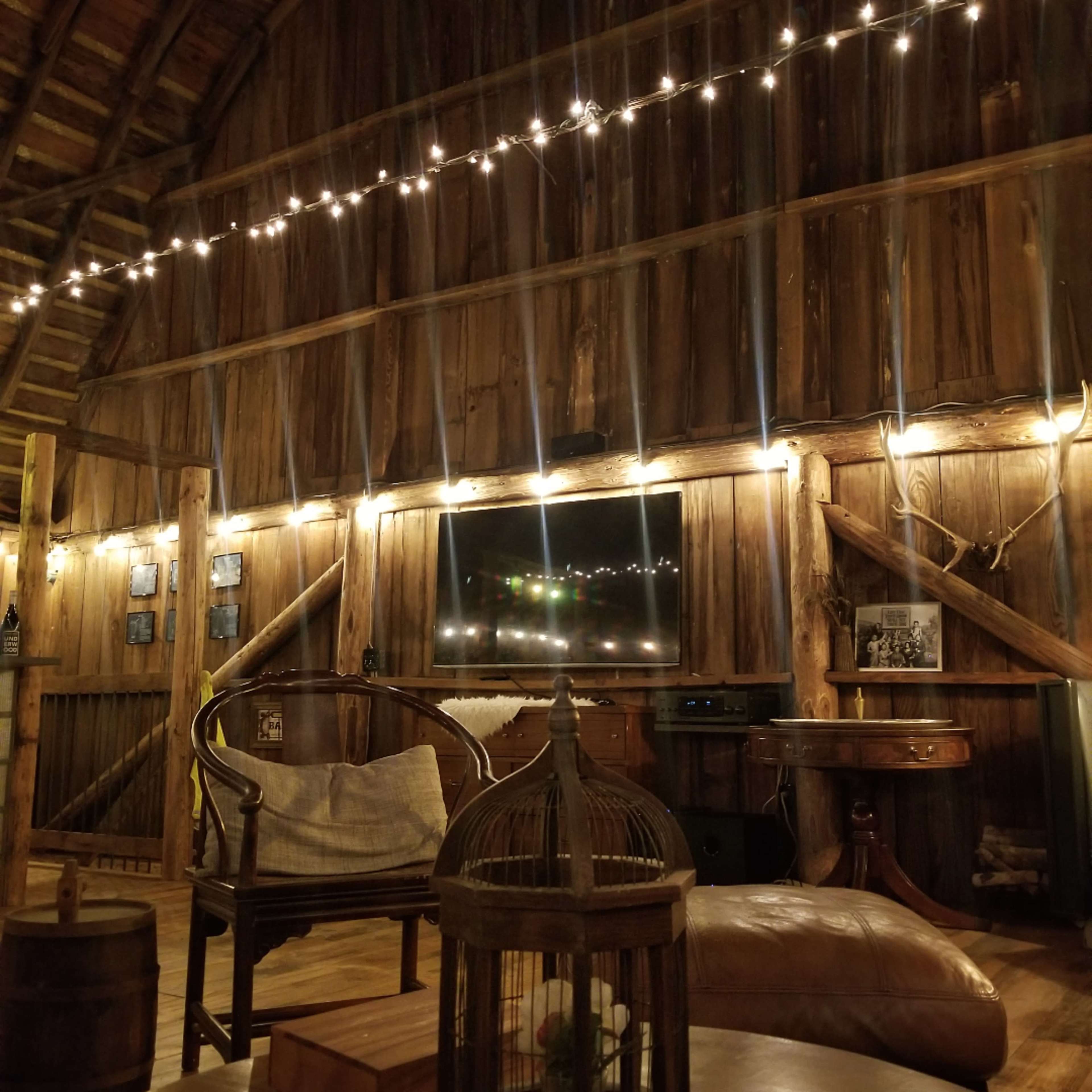 The interior of a rustic barn features wooden walls, string lights, a television, and various seating arrangements.