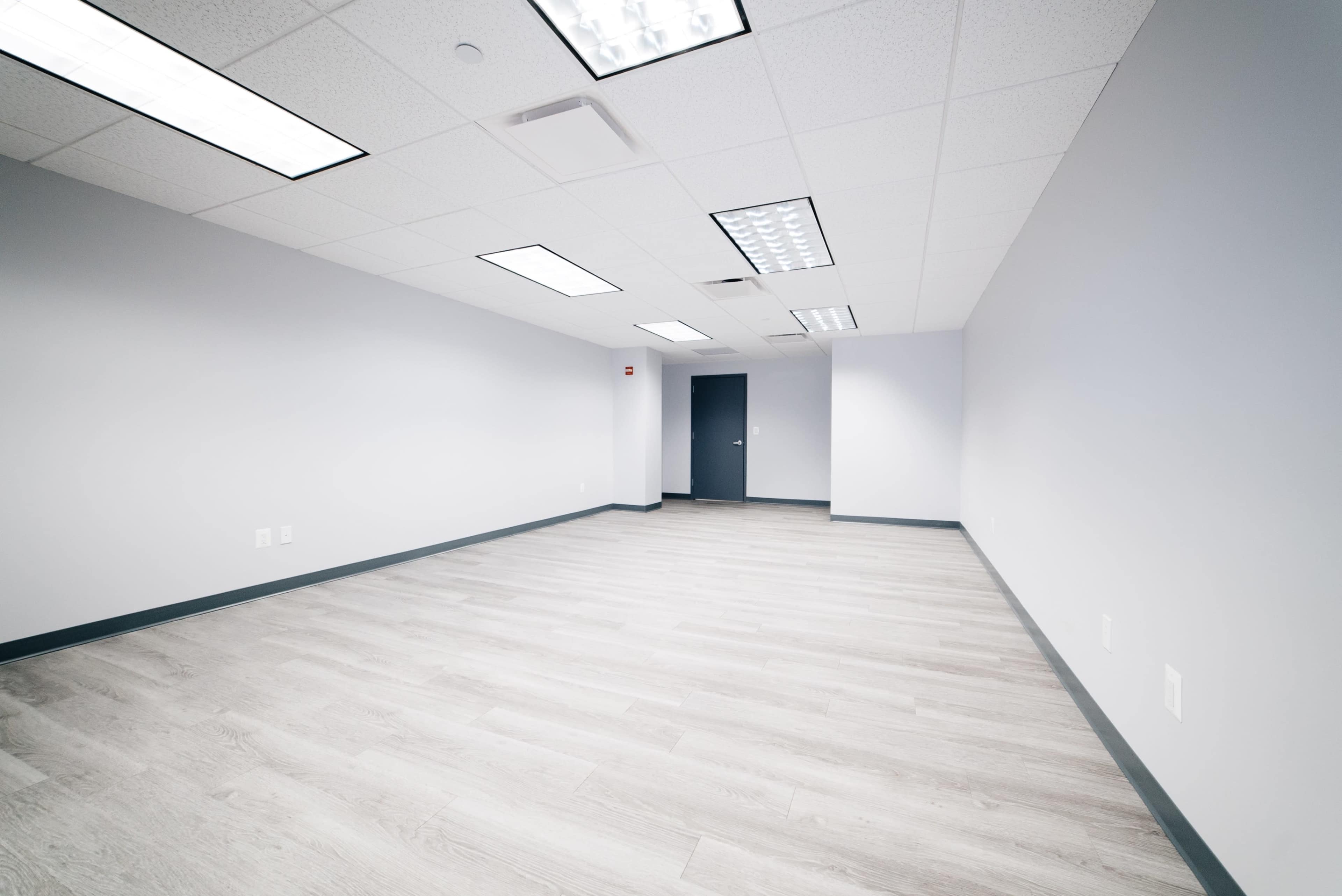 An empty office space with light-colored flooring and gray walls, featuring a single door on one side.