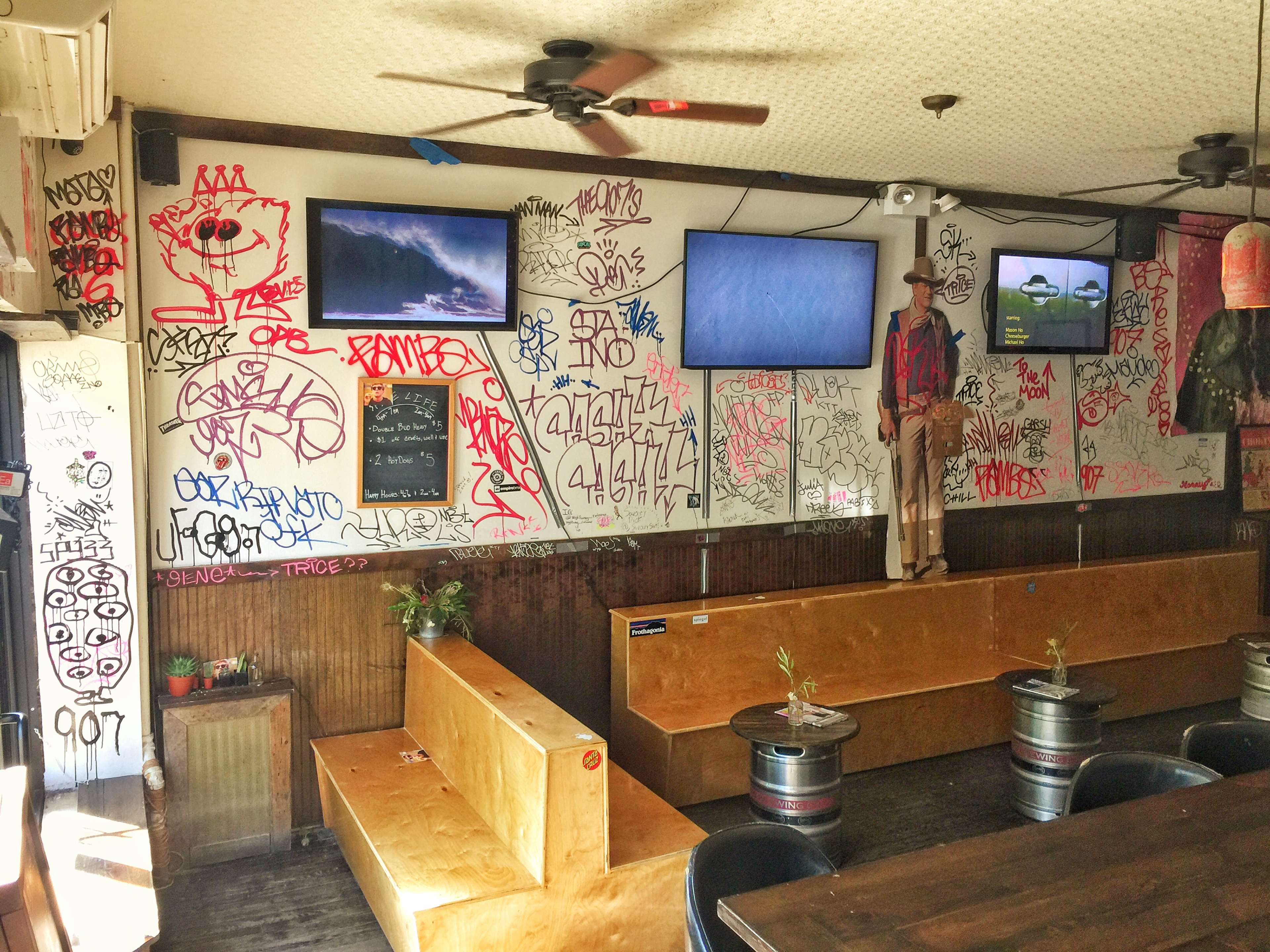 The interior of a bar features walls covered in graffiti, wooden seating areas, and two television screens mounted on the wall.