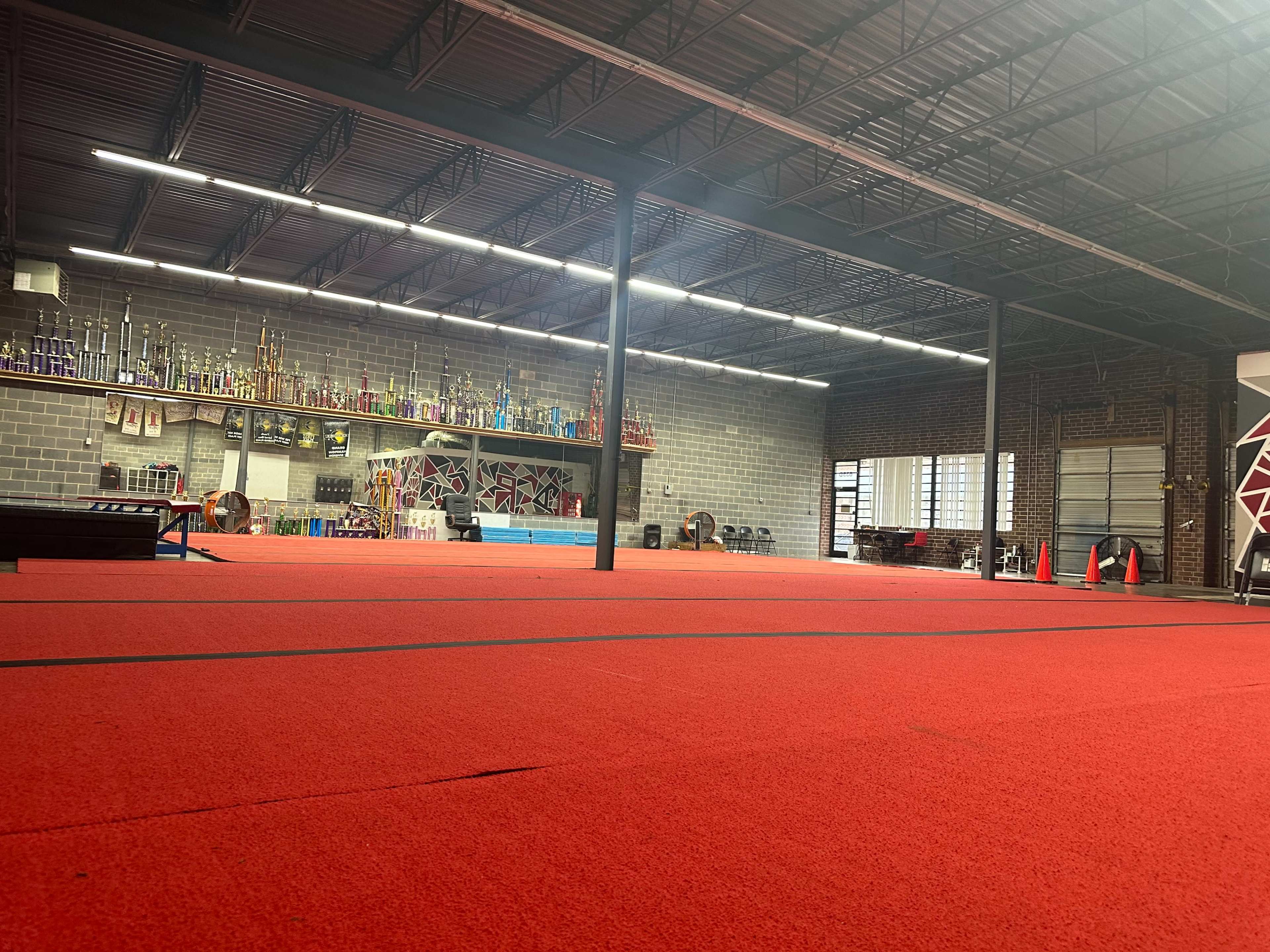 The image shows a gymnasium with red flooring, trophy displays along the back wall, and various gym equipment arranged throughout the space.