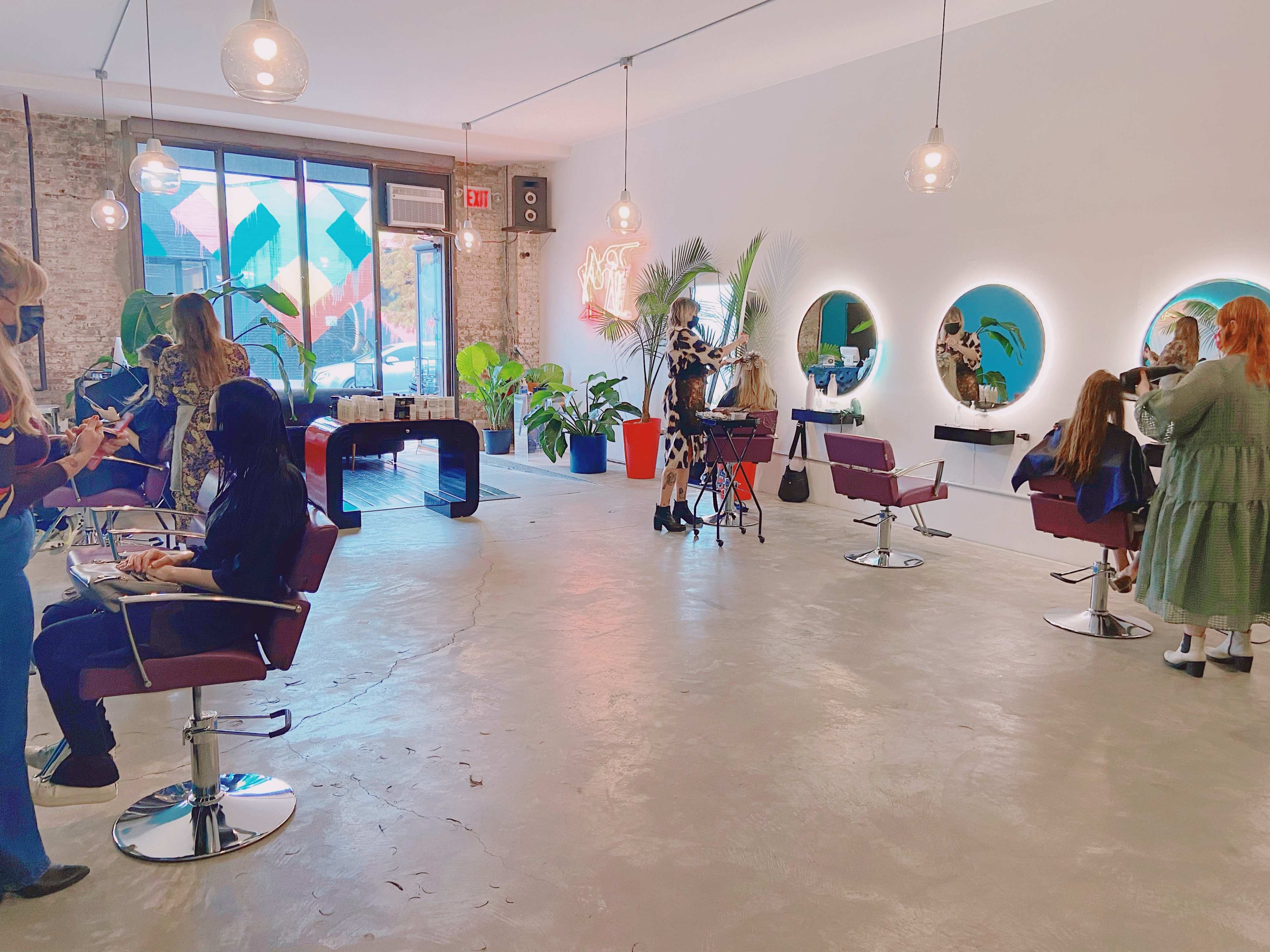 A bright salon interior with several stylists working on clients while surrounded by plants and decorative mirrors.