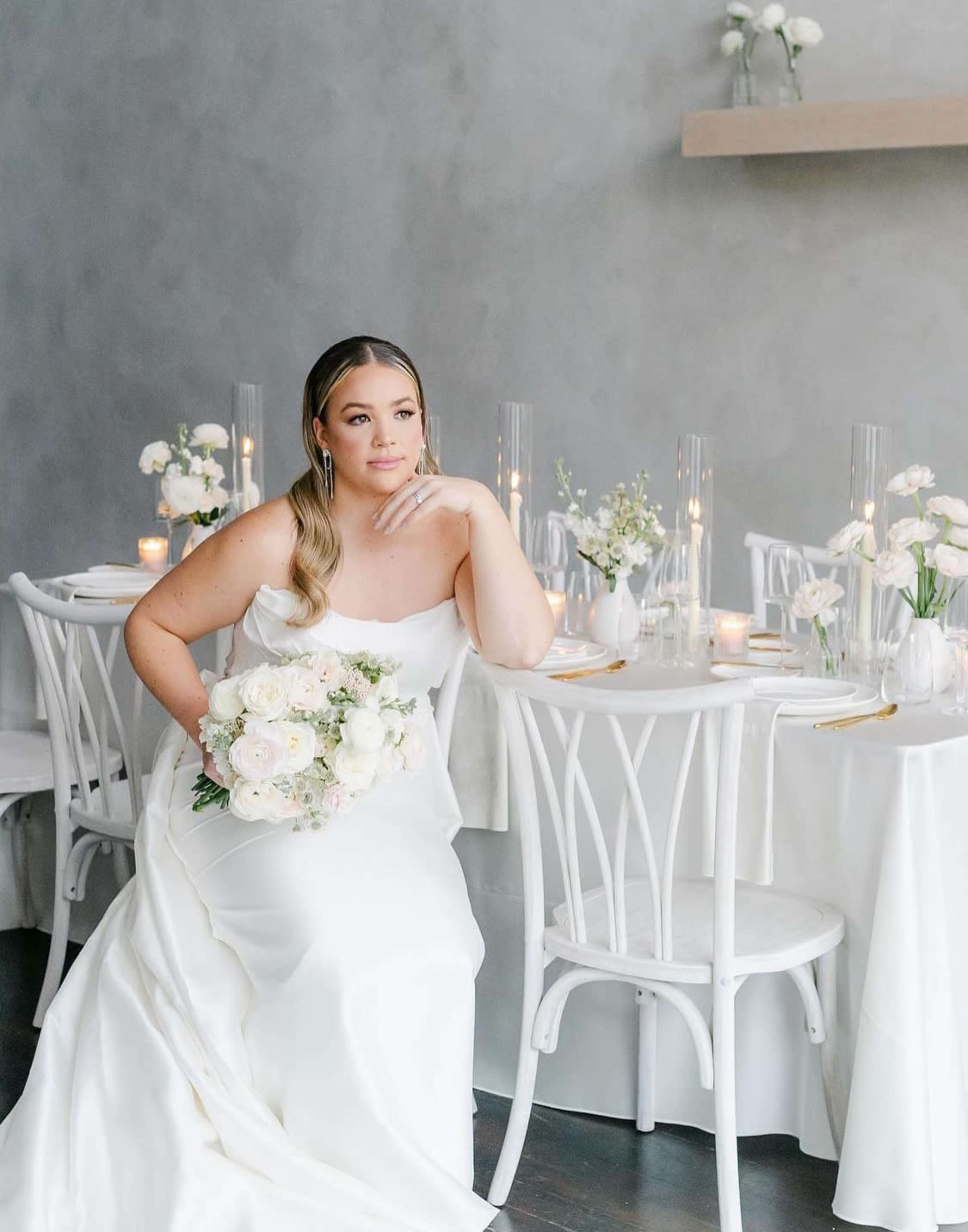 A woman in a white wedding dress sits while holding a bouquet of flowers, surrounded by elegantly set tables adorned with candles and floral arrangements.
