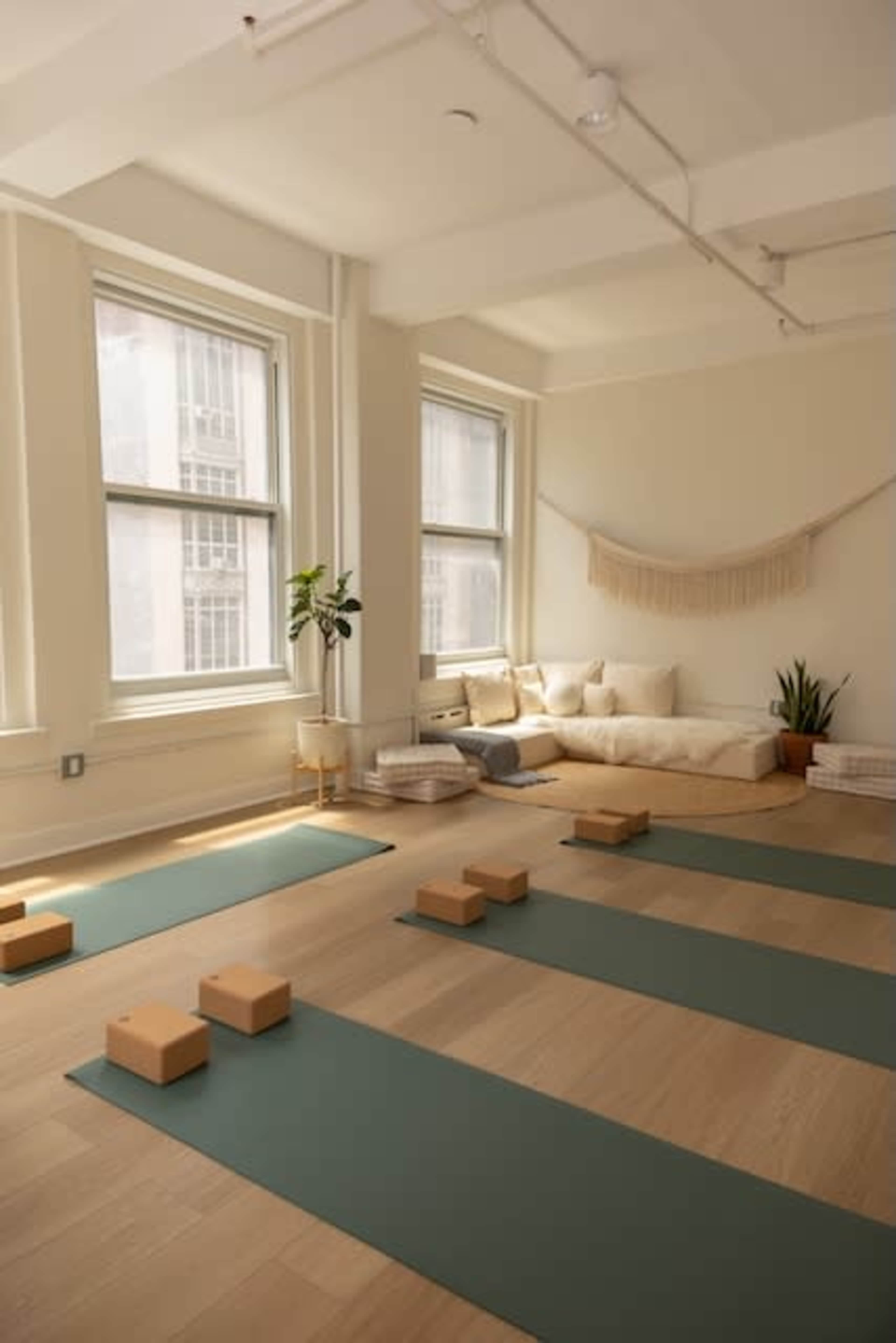 A spacious yoga studio features green mats arranged on a wooden floor, with natural light streaming through large windows and a cozy seating area in the background.