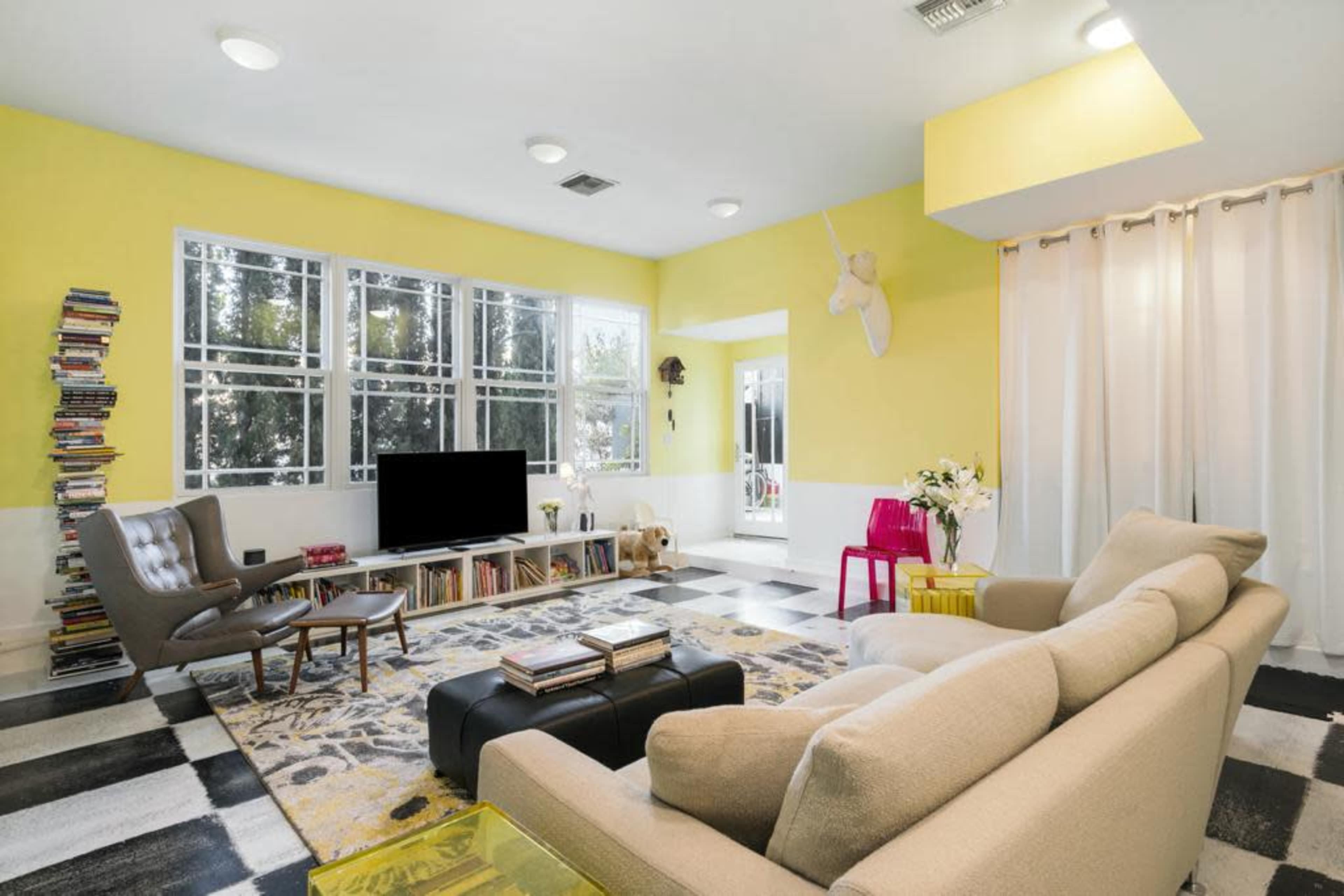 A bright living room features a yellow accent wall, a large television, modern furniture, and a wall of arranged books.