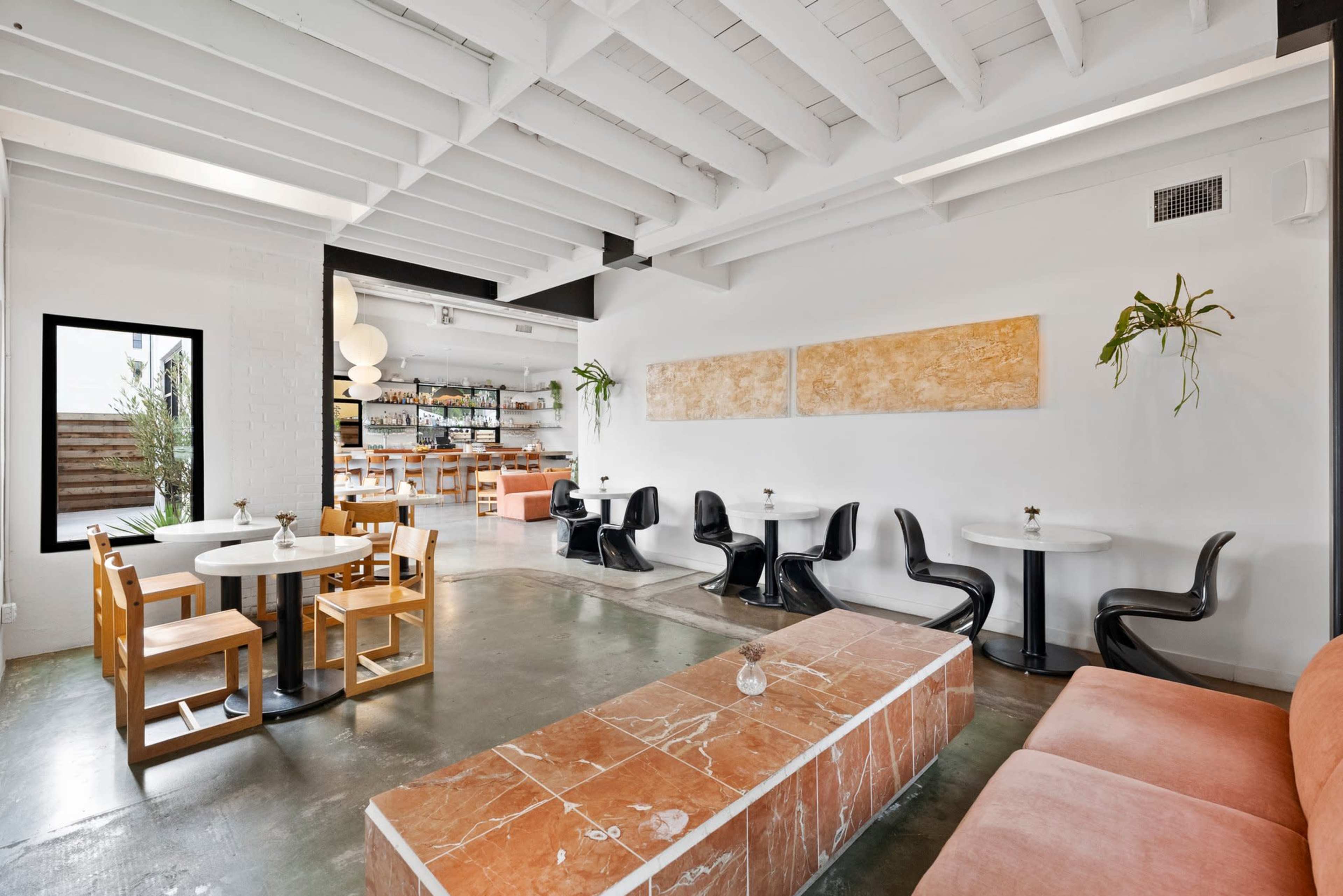 The image shows a modern café interior featuring circular tables, black chairs, a long marble bench, and bright white walls with minimal decor.