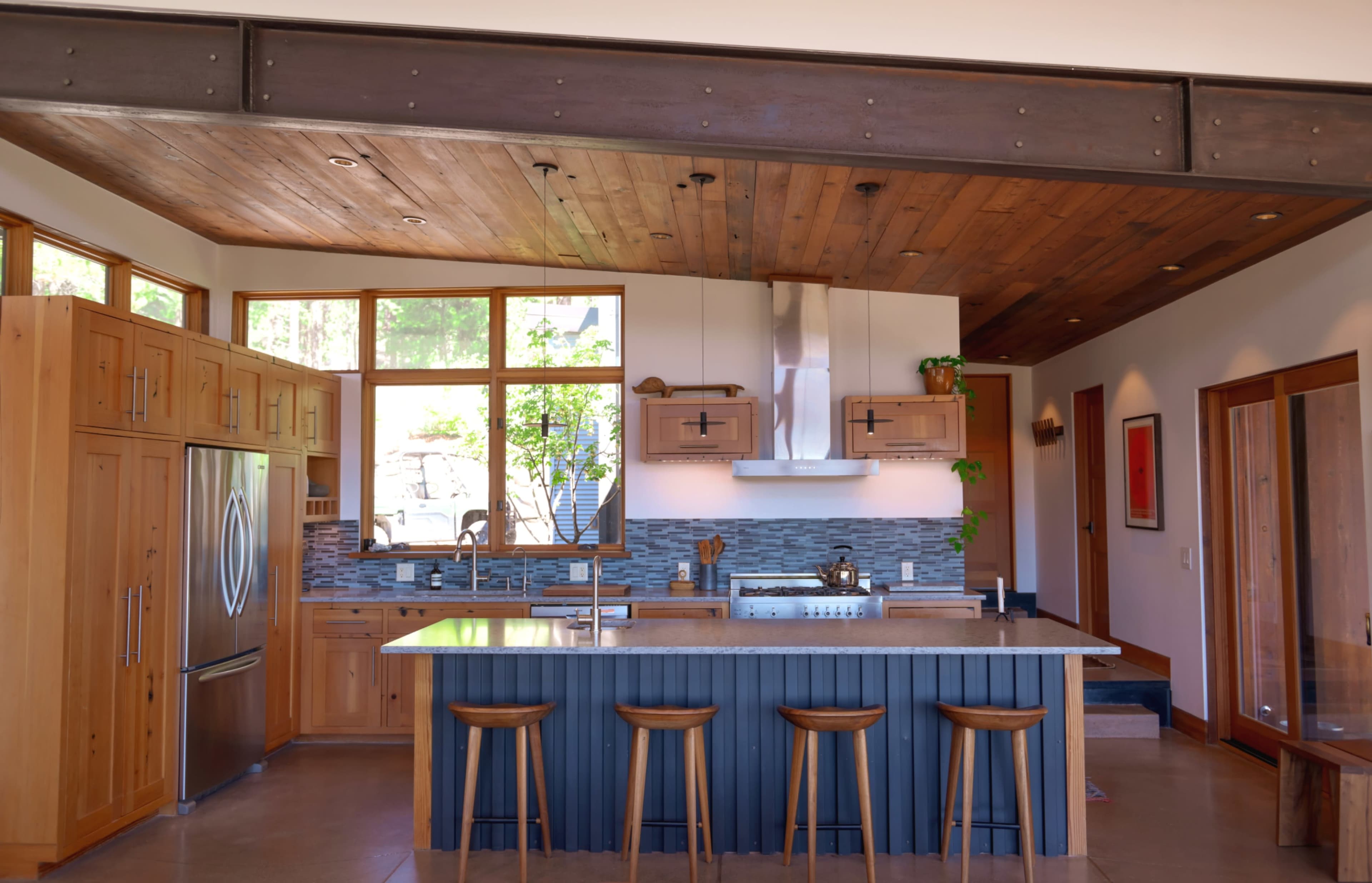 A modern kitchen features wooden cabinets, a large island with bar stools, and a stainless steel cooktop beneath a hood.