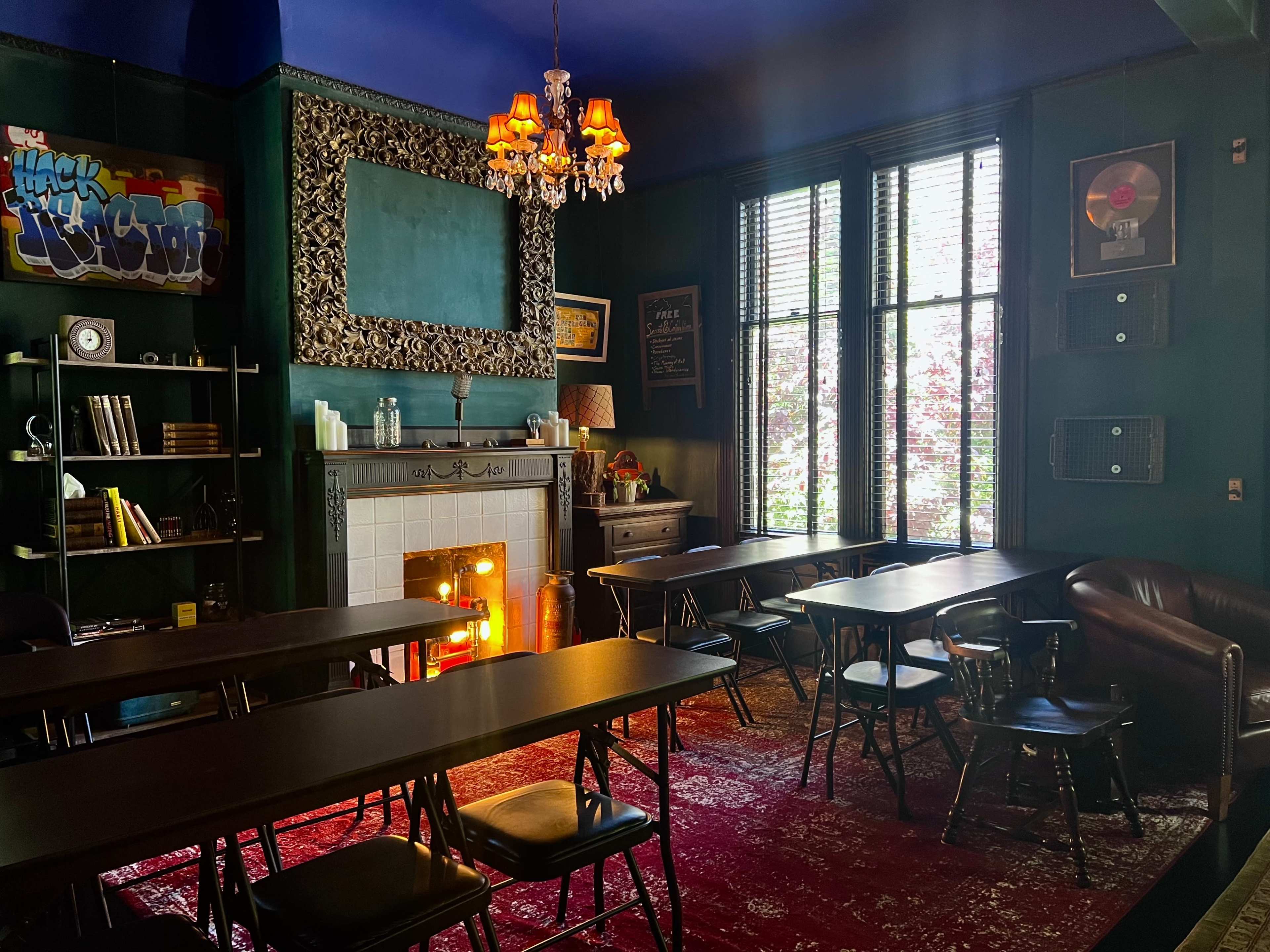 A dimly lit room with rows of black folding tables, a decorative fireplace, and large windows overlooking greenery.