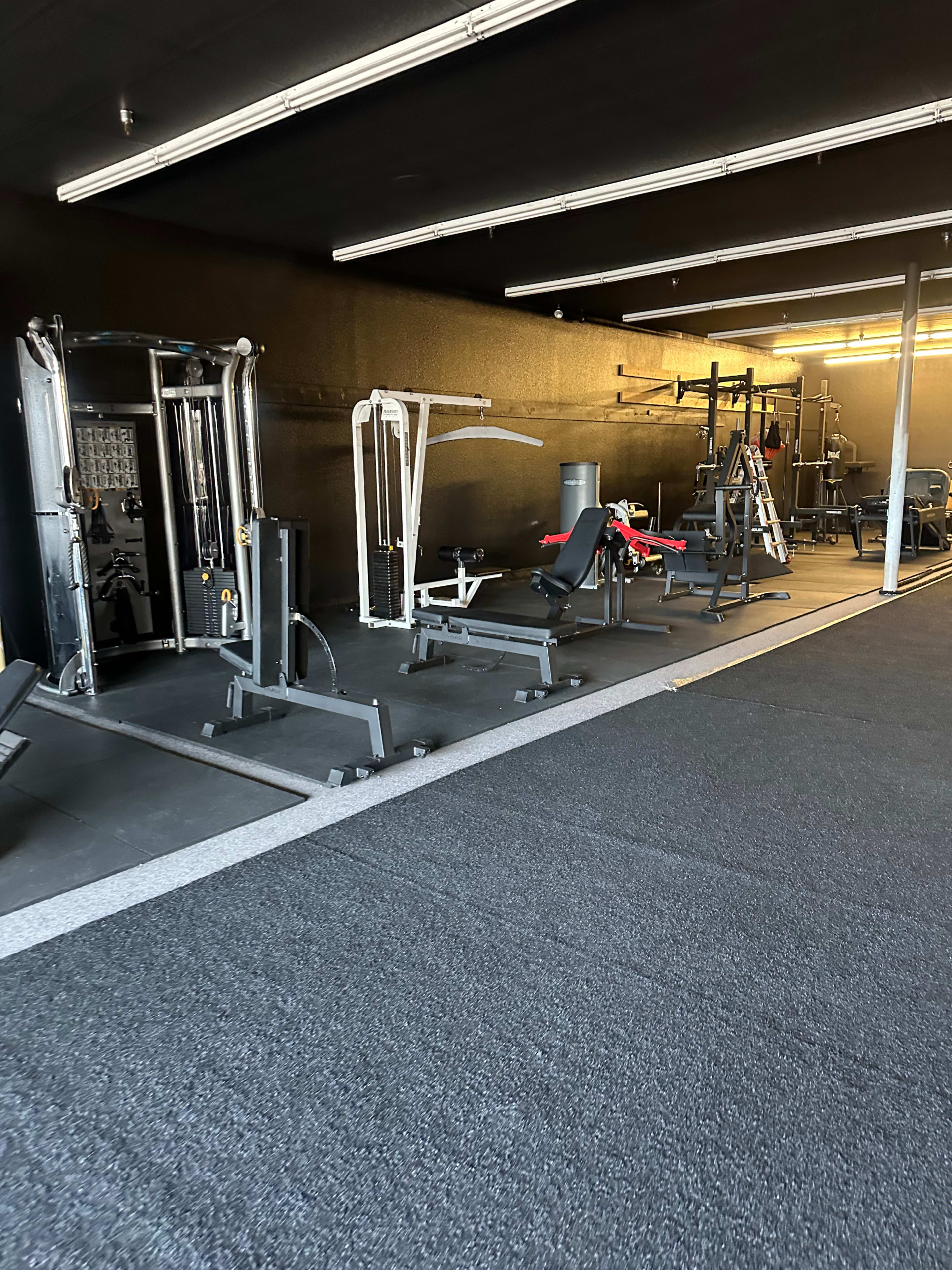 The image shows a gym interior featuring various exercise equipment arranged in a spacious layout with dark walls and flooring.