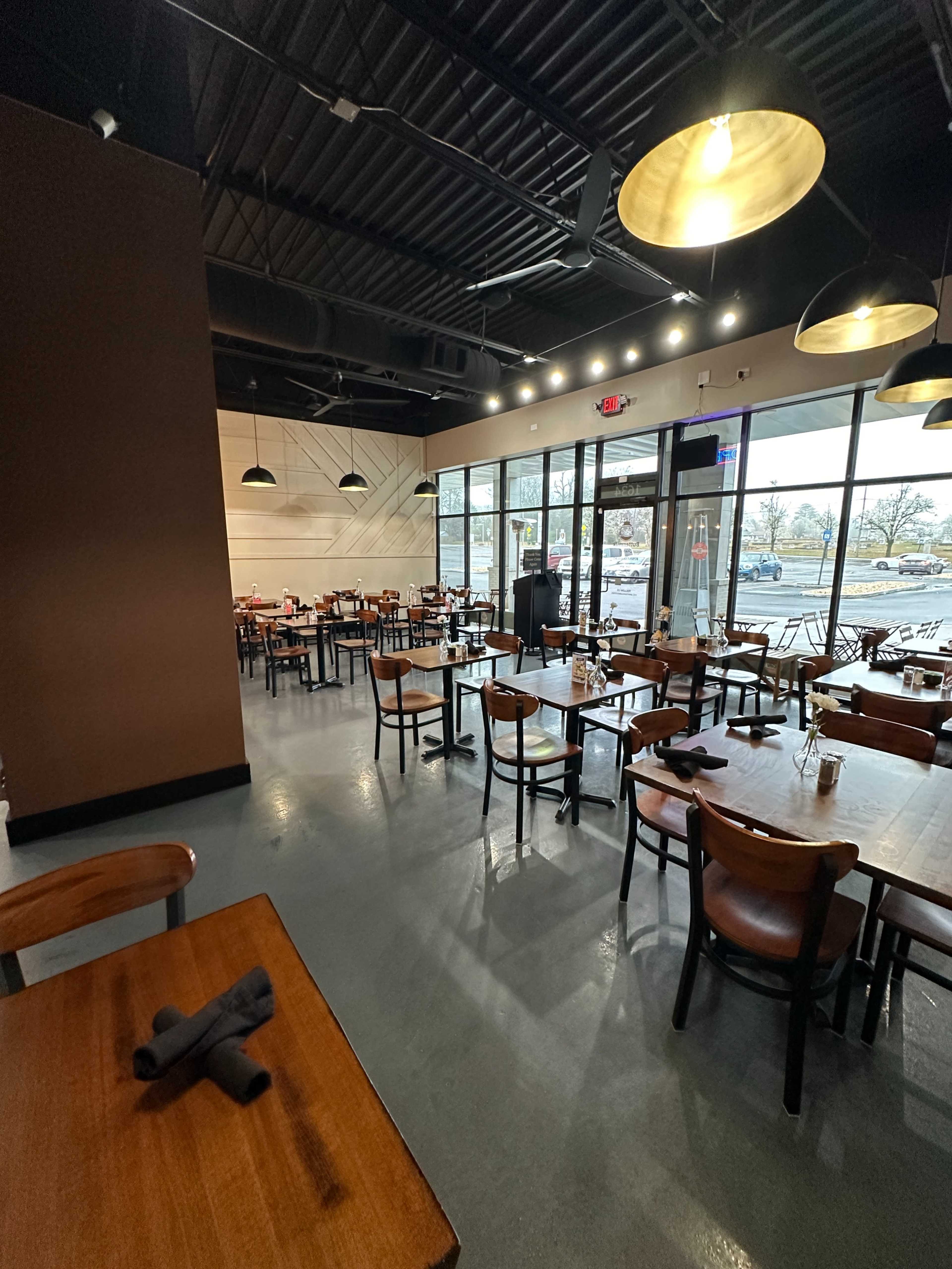 The interior of a modern restaurant with empty tables and chairs arranged neatly, illuminated by overhead lights and large windows.