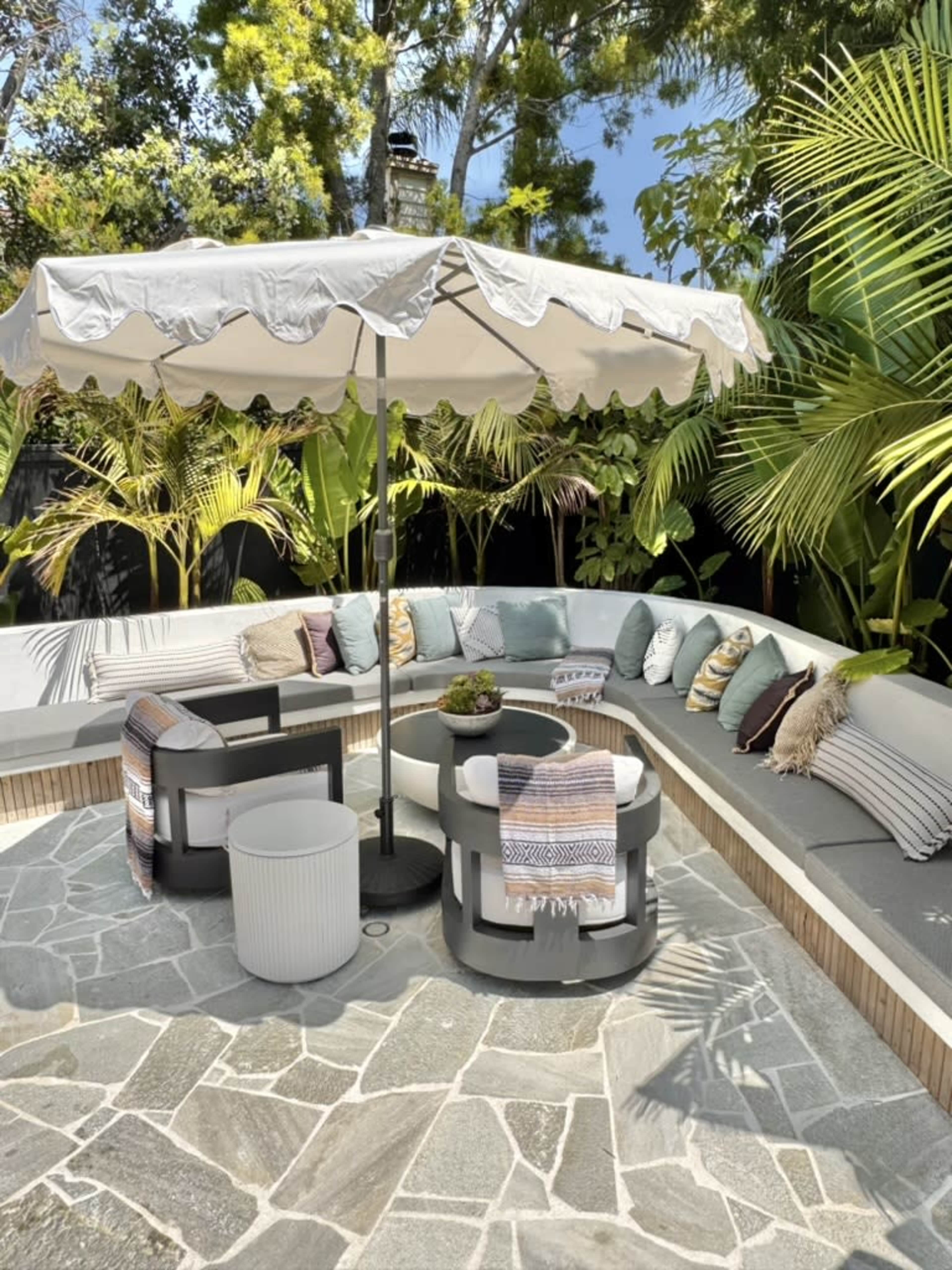 A circular seating area with a large umbrella is surrounded by lush greenery, featuring cushions, throws, and a small table with a centerpiece.
