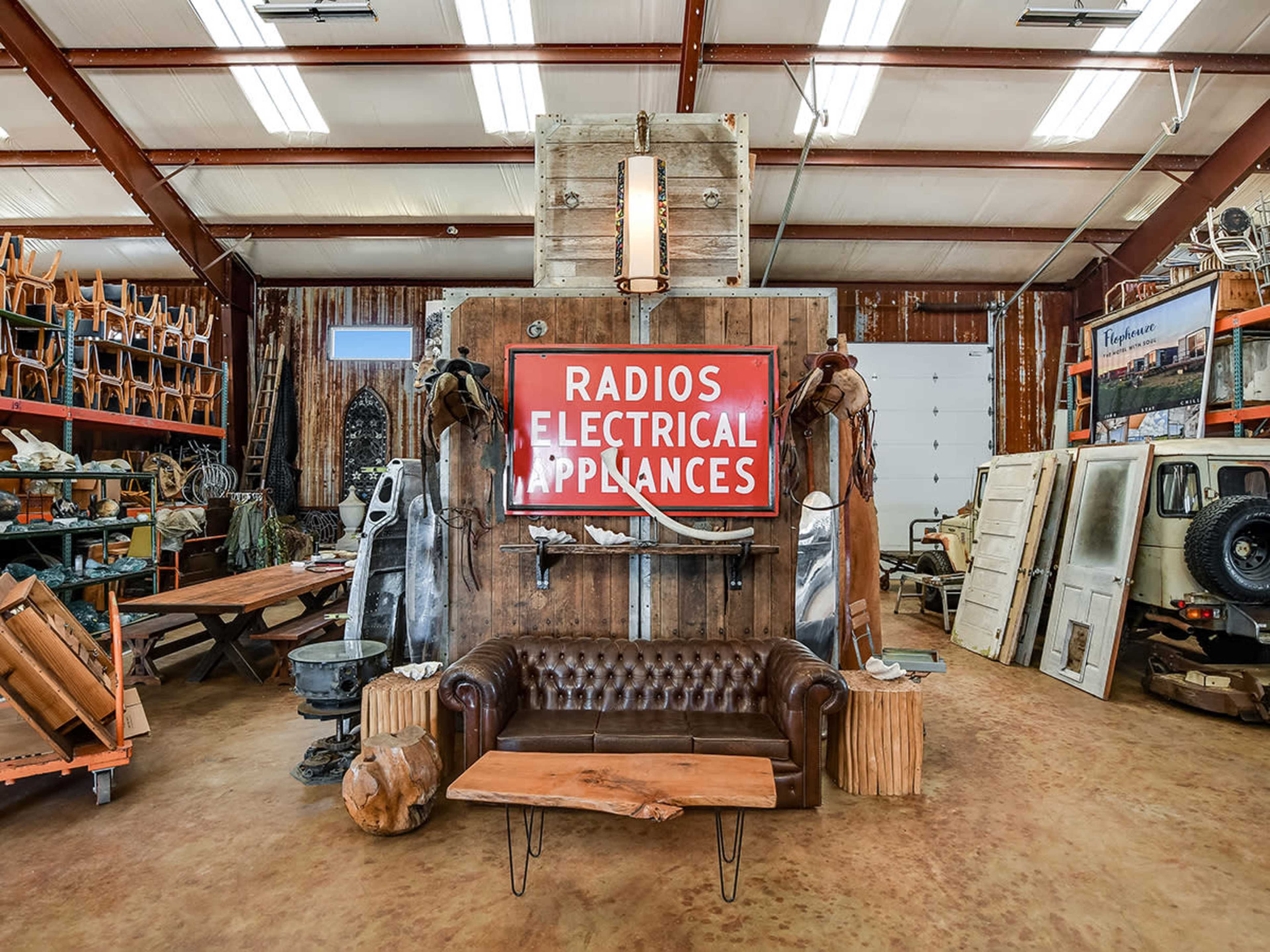 The interior of a rustic shop features a vintage leather sofa in front of a large sign that reads "RADIOS ELECTRICAL APPLIANCES" against a backdrop of reclaimed wood and various displayed items.