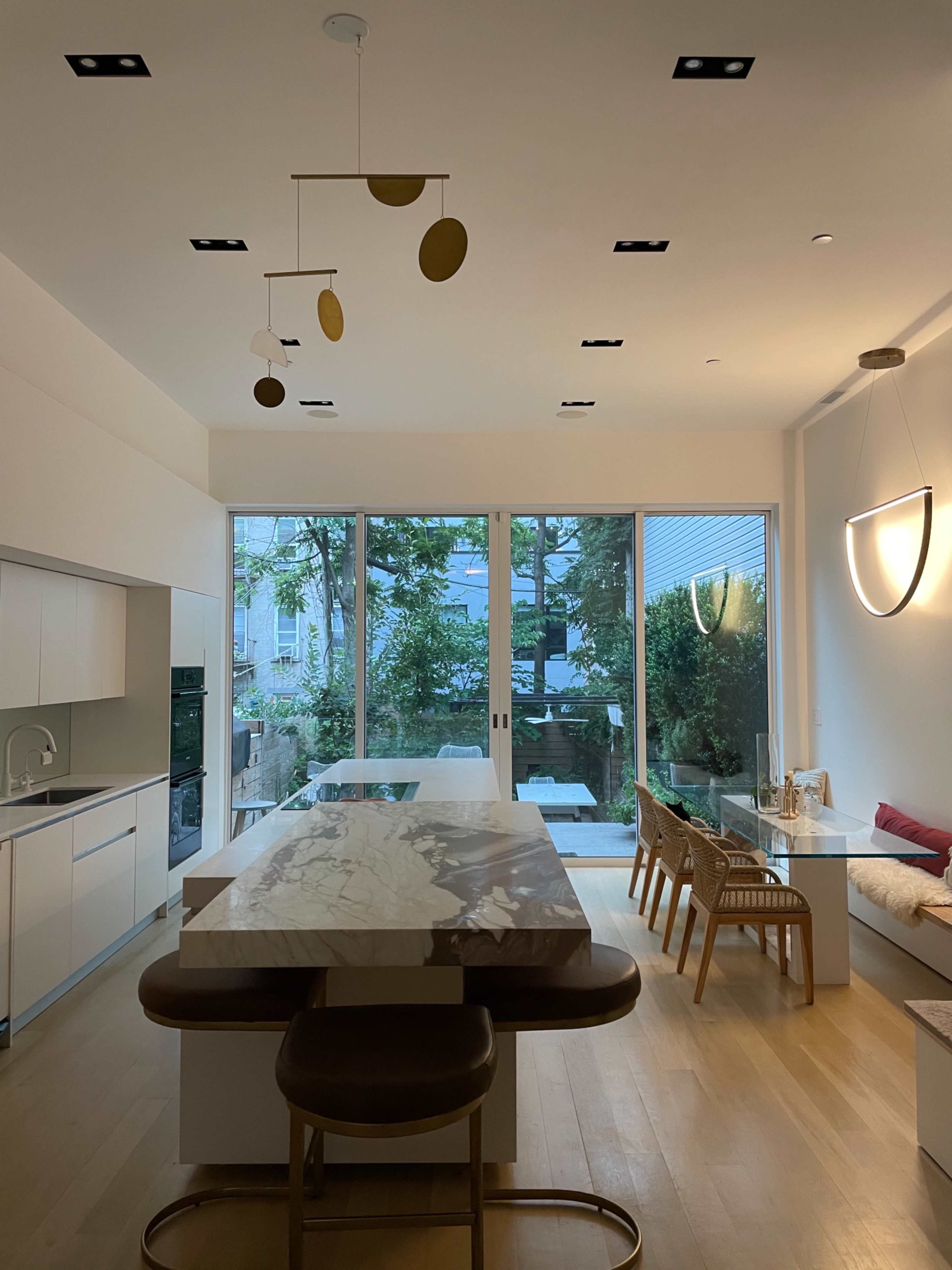 A modern kitchen features a large marble island, minimalist cabinetry, and large windows overlooking a garden.