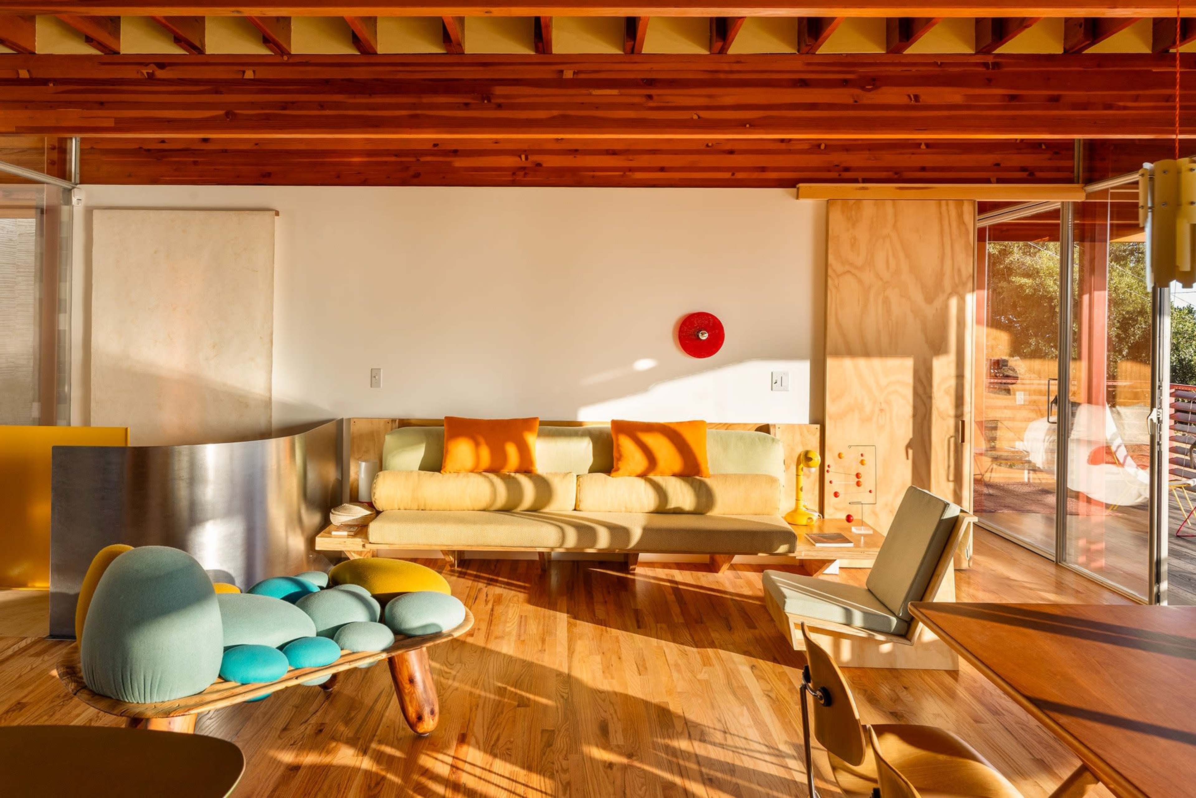 The image shows a modern interior living space with wooden beams, a yellow couch, colorful seating, and large glass doors that open to a patio.