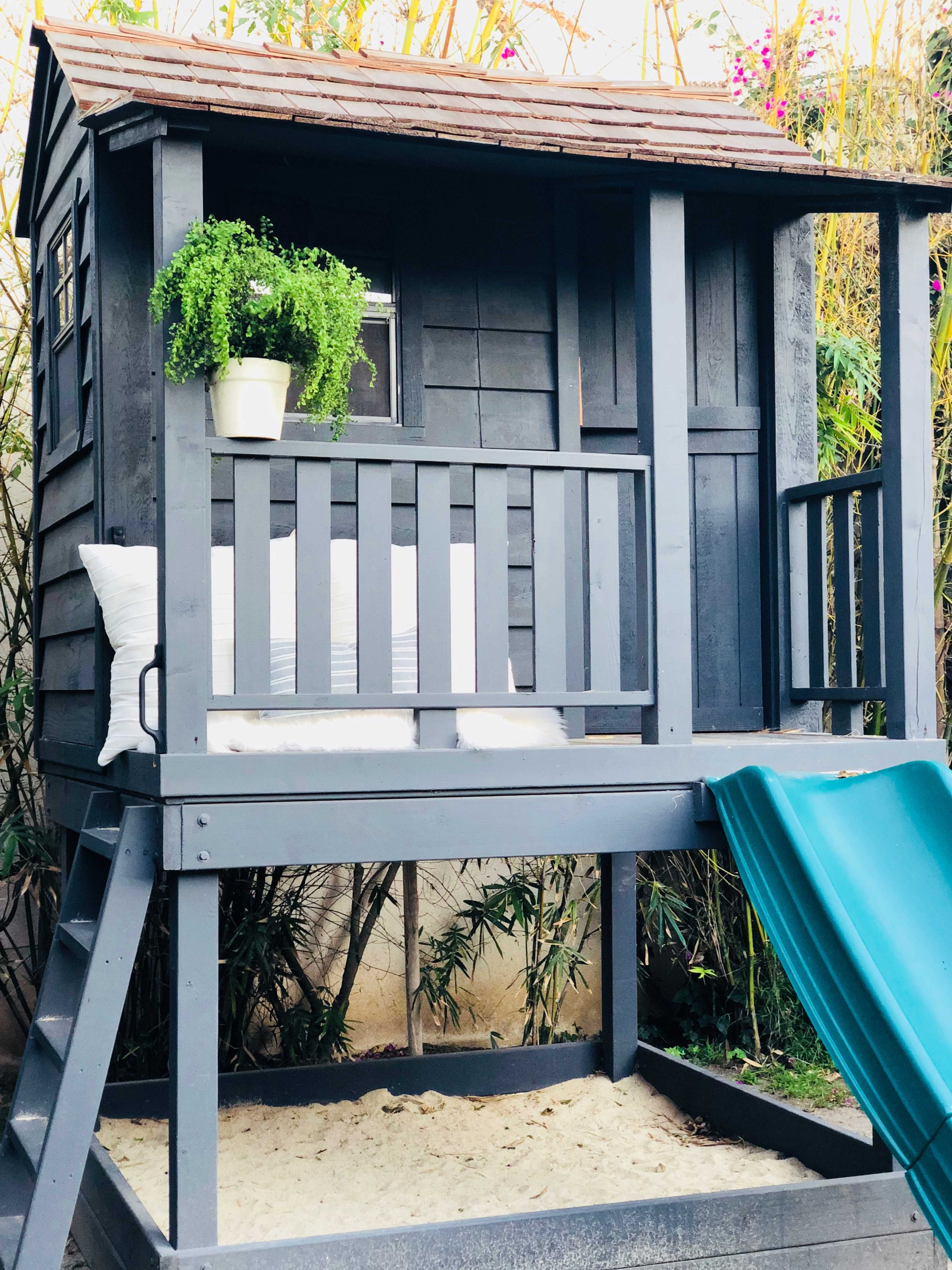 A wooden playhouse elevated on posts, featuring a slide, planter, and seating on the porch.