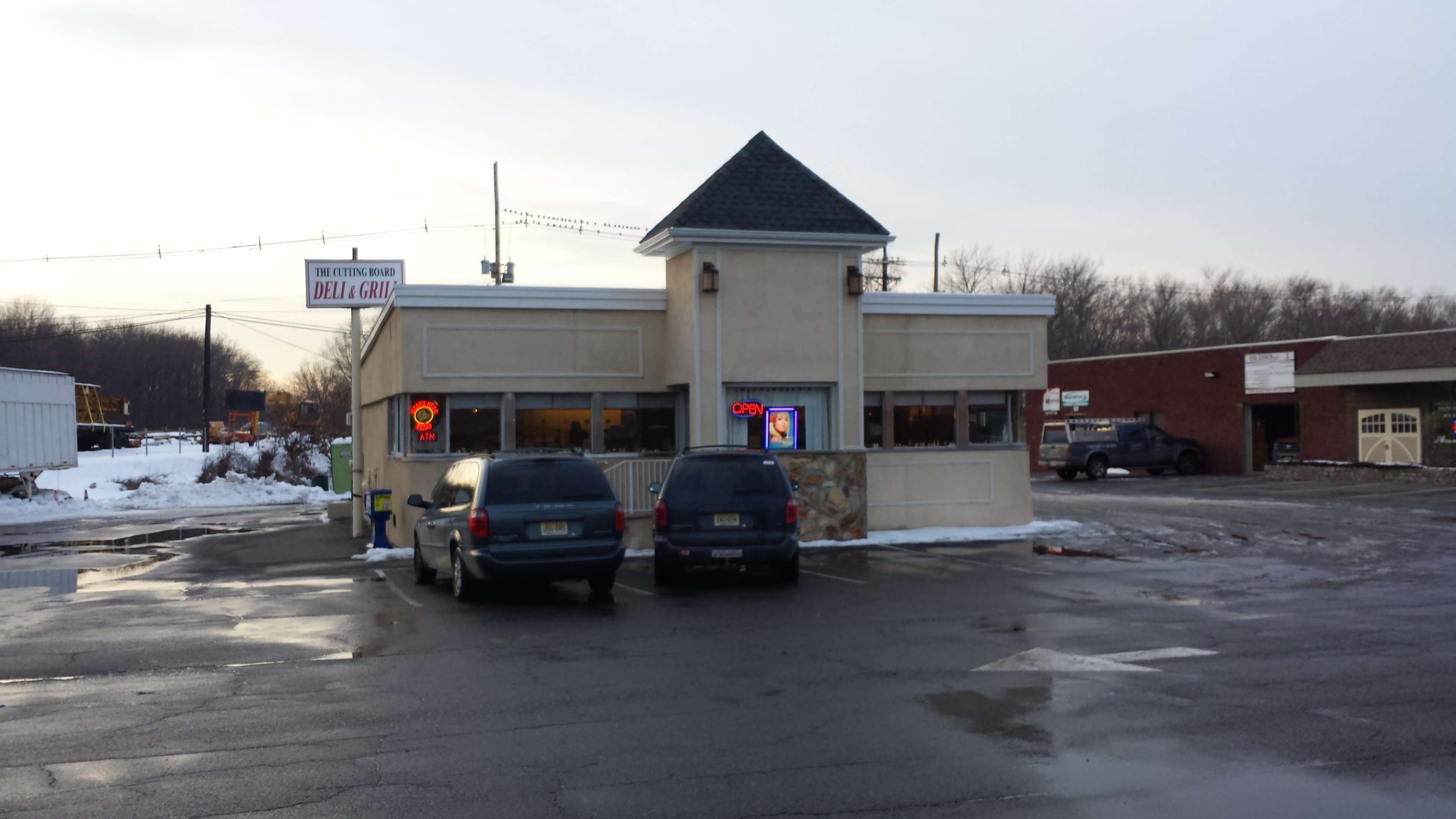 A small deli with a peaked roof has large windows and a parking area in front, featuring two parked vans.