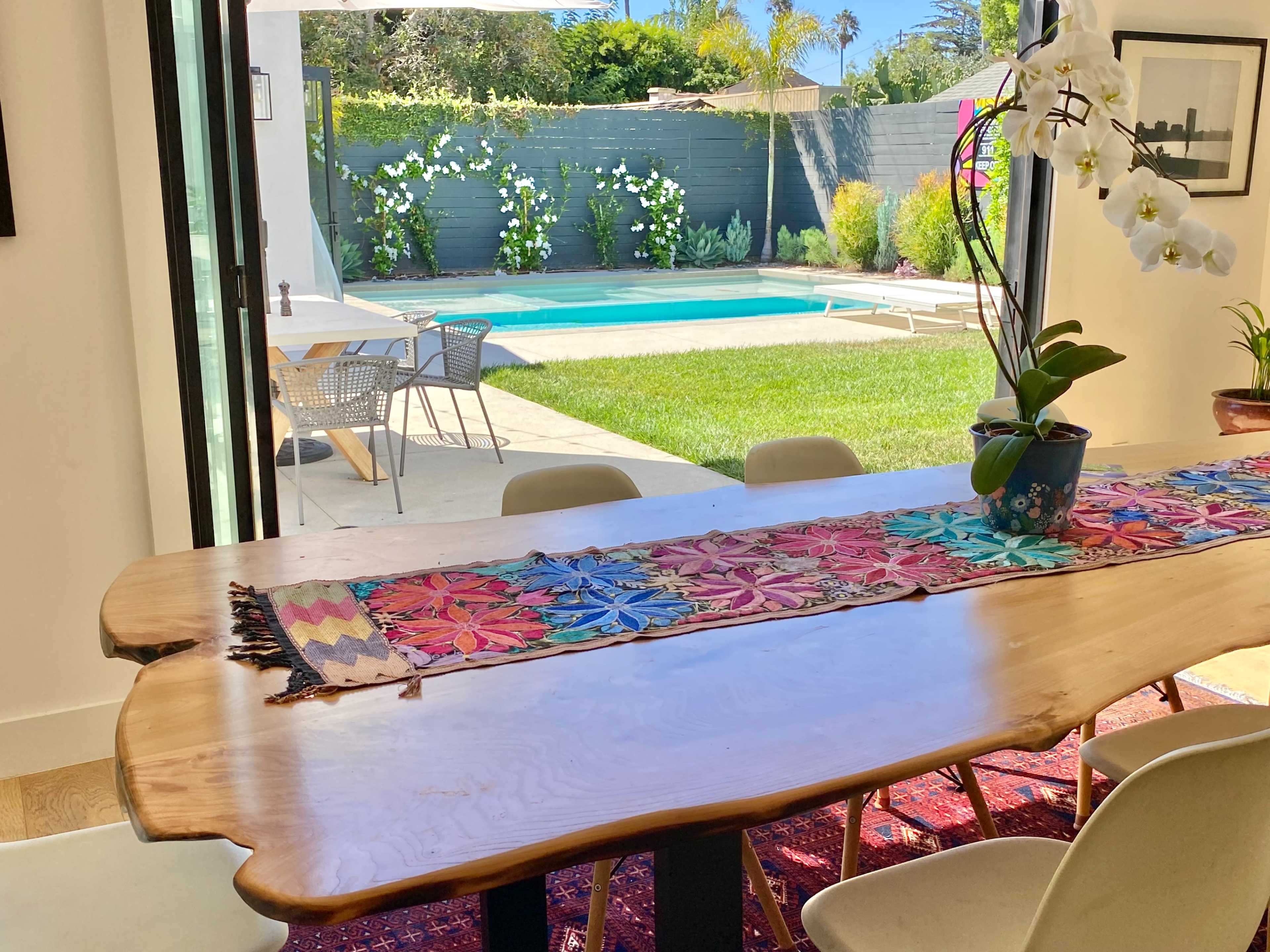 The scene features a wooden dining table adorned with a colorful runner, overlooking a backyard with a swimming pool and landscaped greenery.