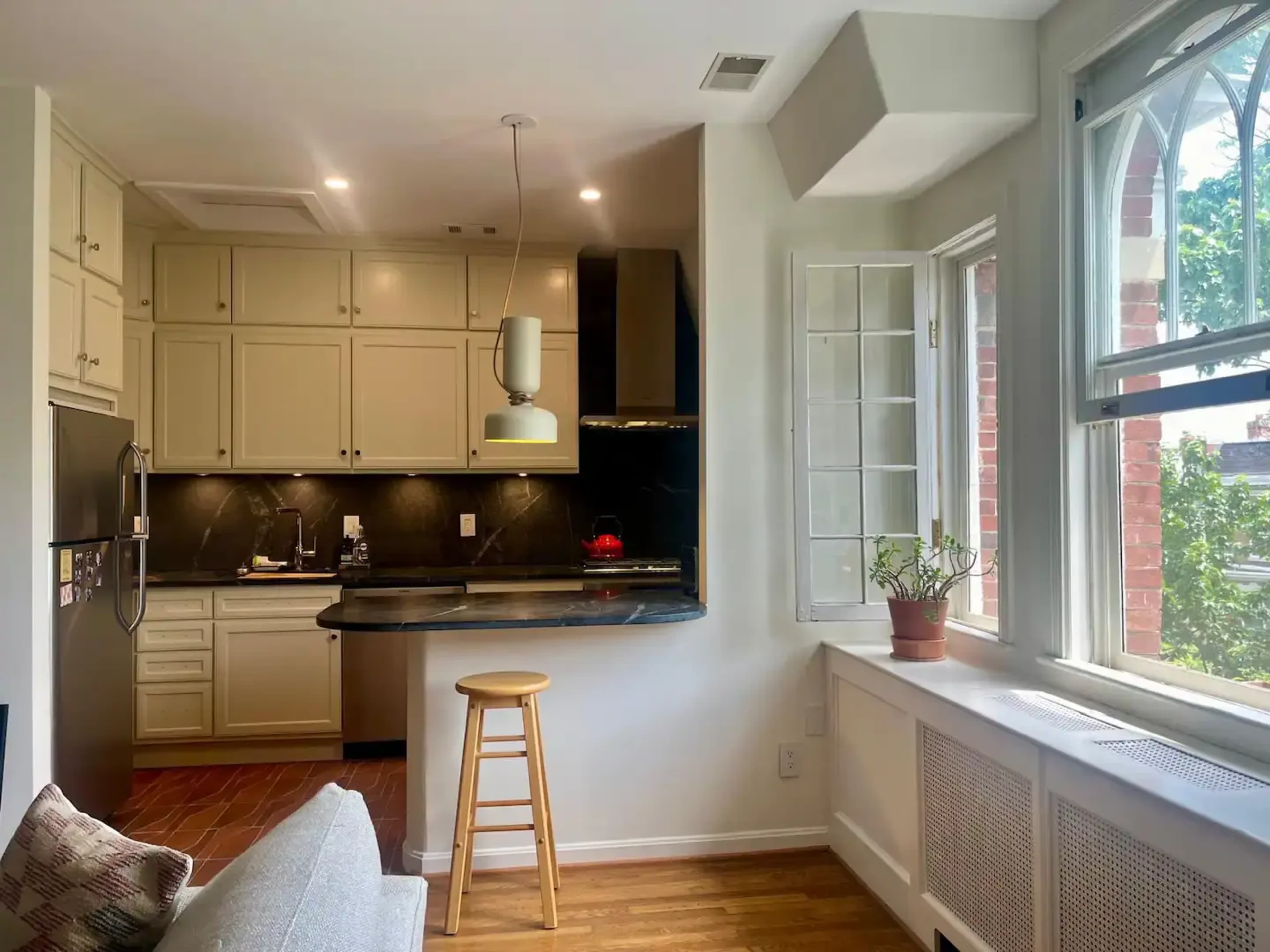 A compact kitchen features light cabinetry, a dark countertop, and a tall window allowing natural light to enter the space.