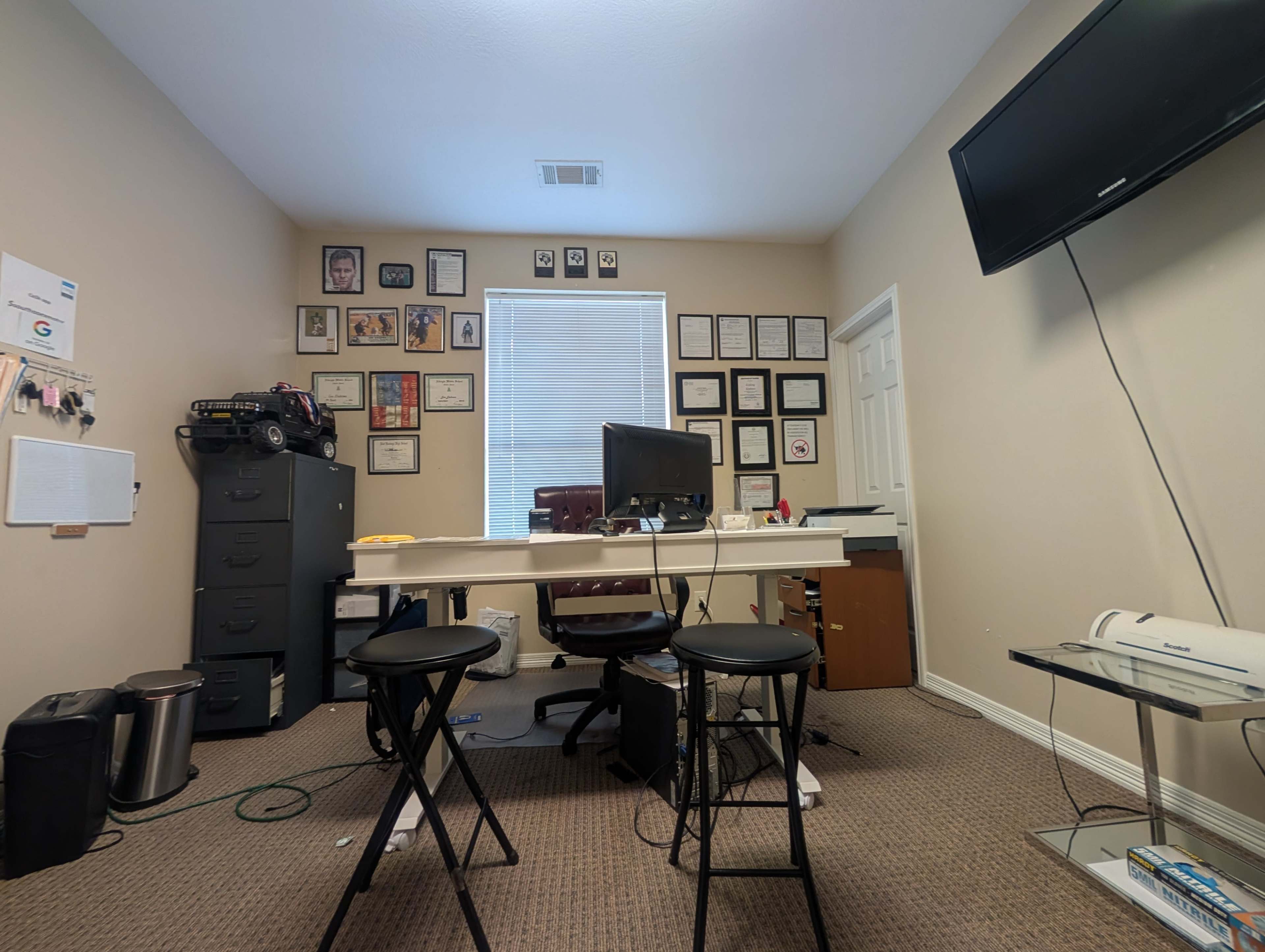 A small office features a desk with a computer, two stools, and a wall adorned with various framed certificates and photographs.