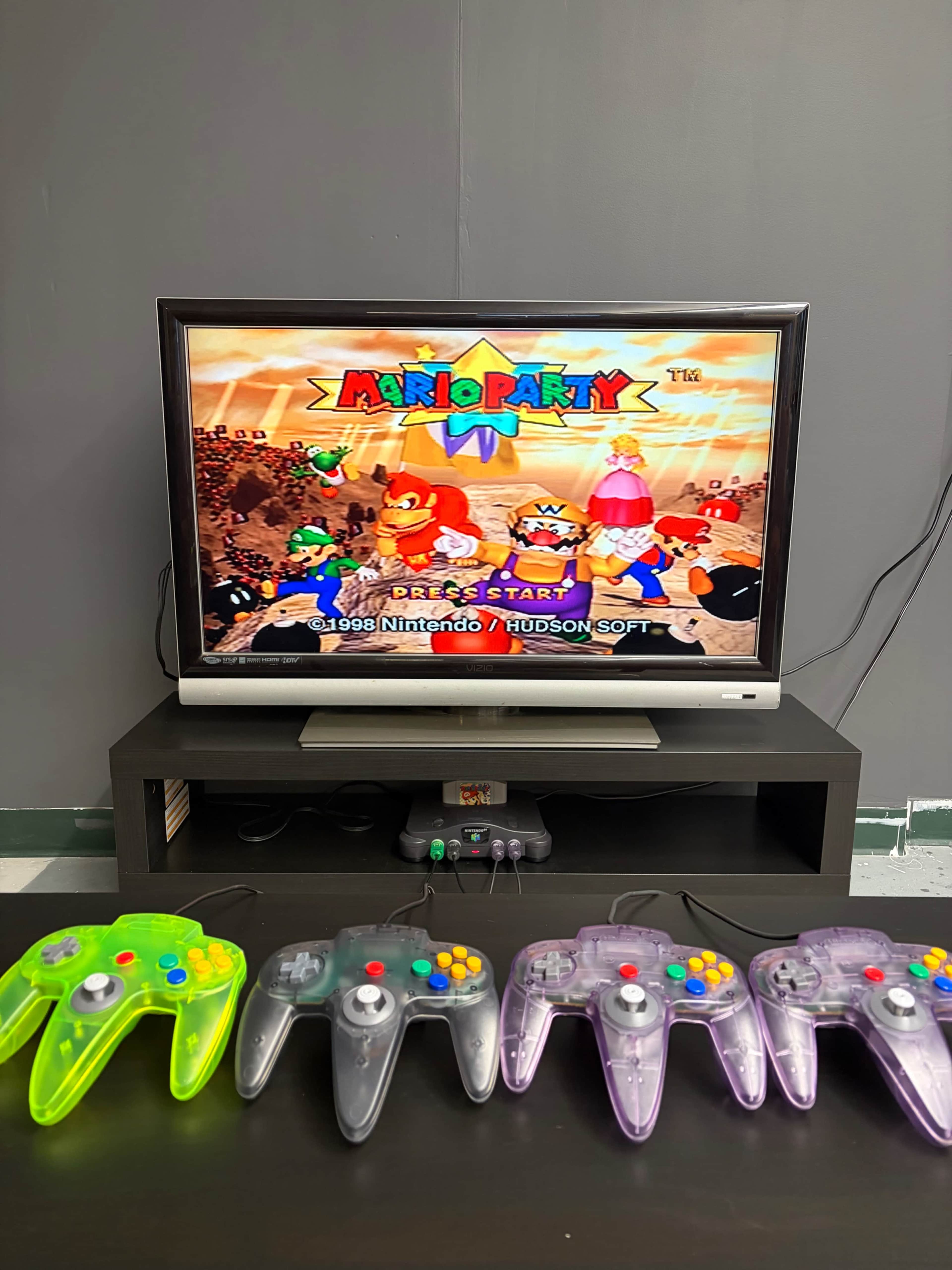 A television displaying the game "Mario Party" is placed on a black entertainment unit, with four Nintendo 64 controllers in various colors positioned in front of it.