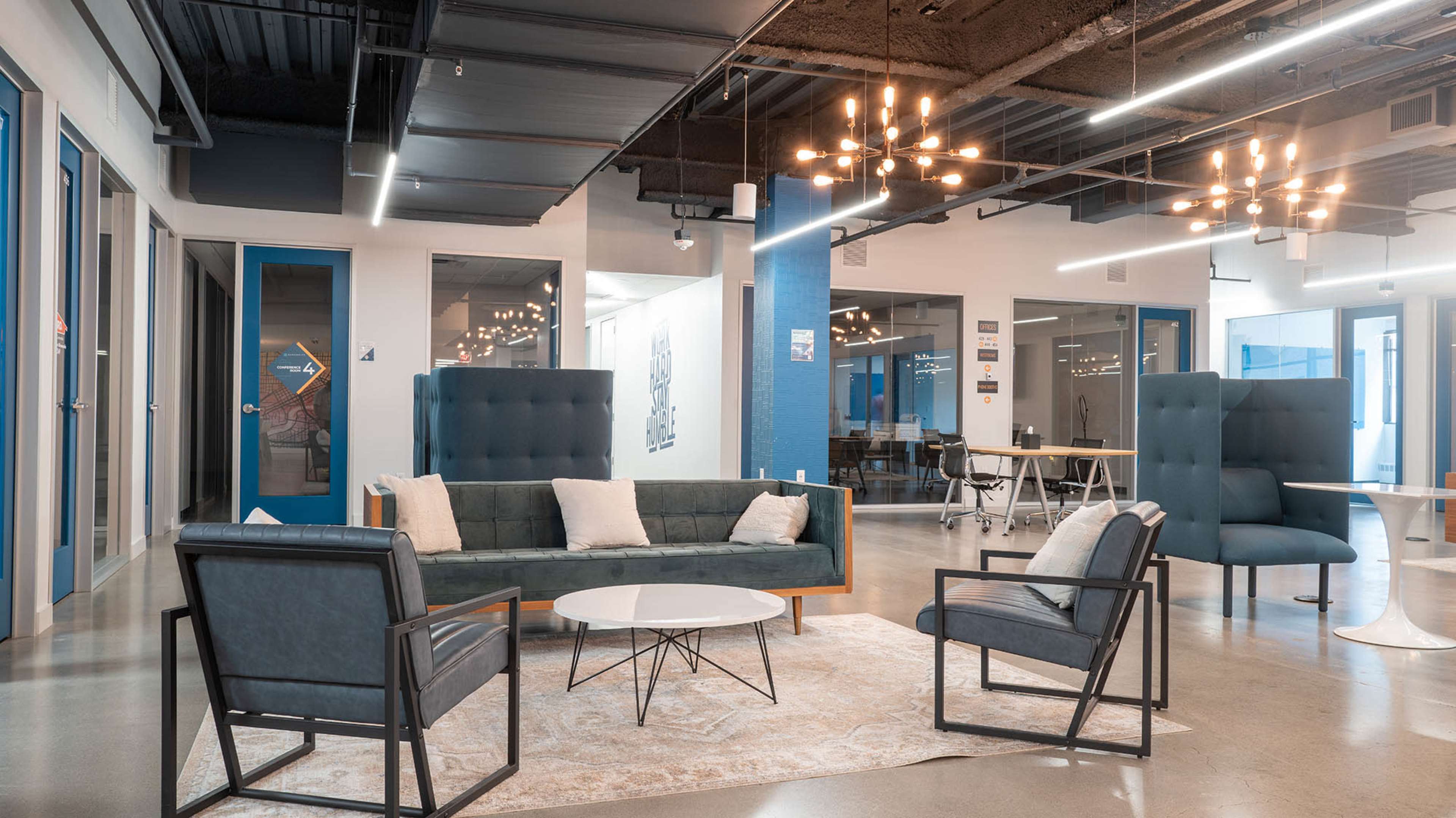 The image shows a modern office lounge area with a green sofa, two black chairs, a circular coffee table, and artistic lighting fixtures.