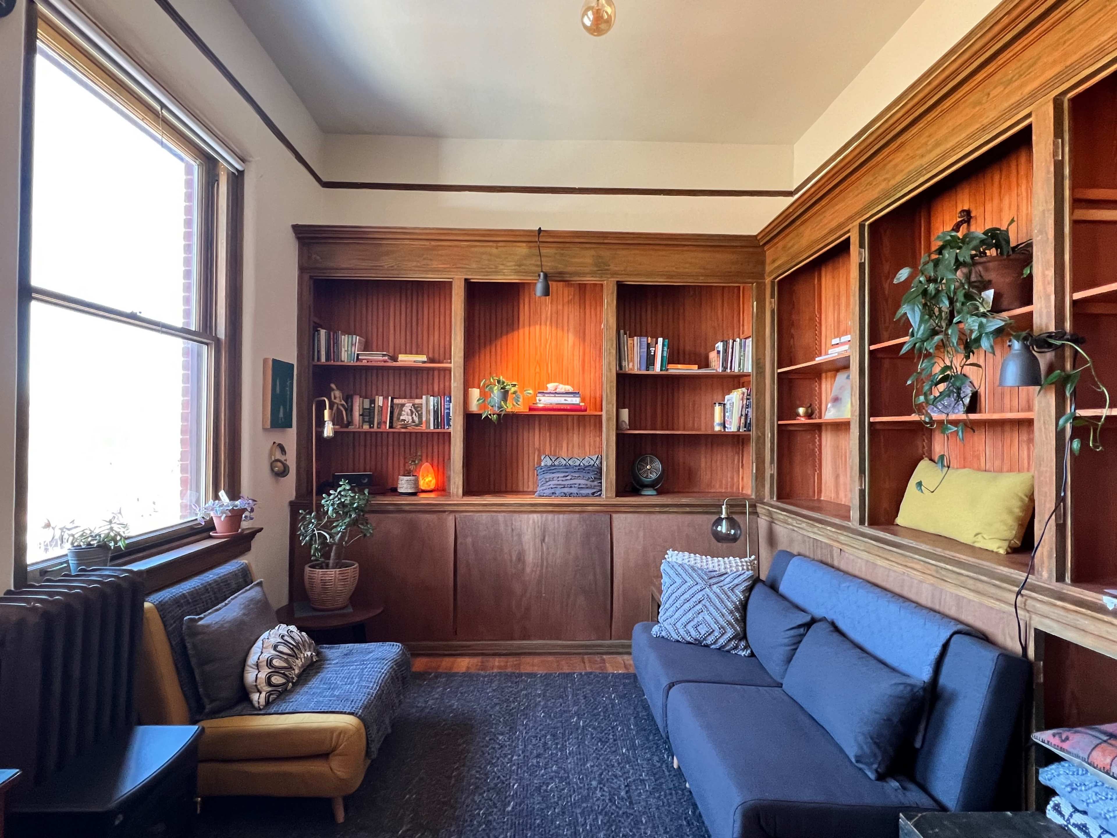 A cozy living room with a dark couch, an armchair, wooden bookshelves filled with books, and a large window letting in natural light.