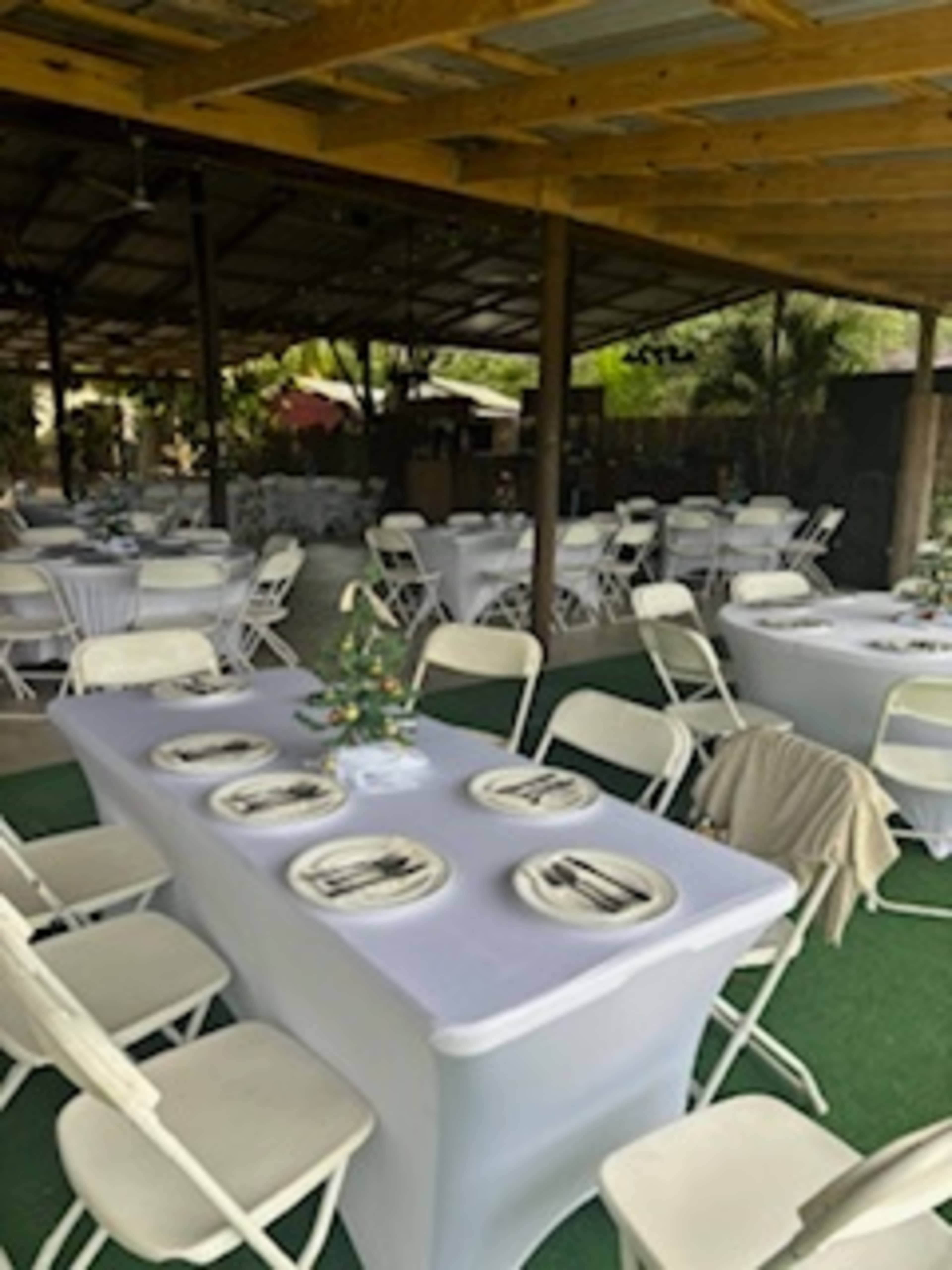 The image shows a banquet setting with several tables covered in white cloth and arranged with plates and cutlery, all under a wooden structure.