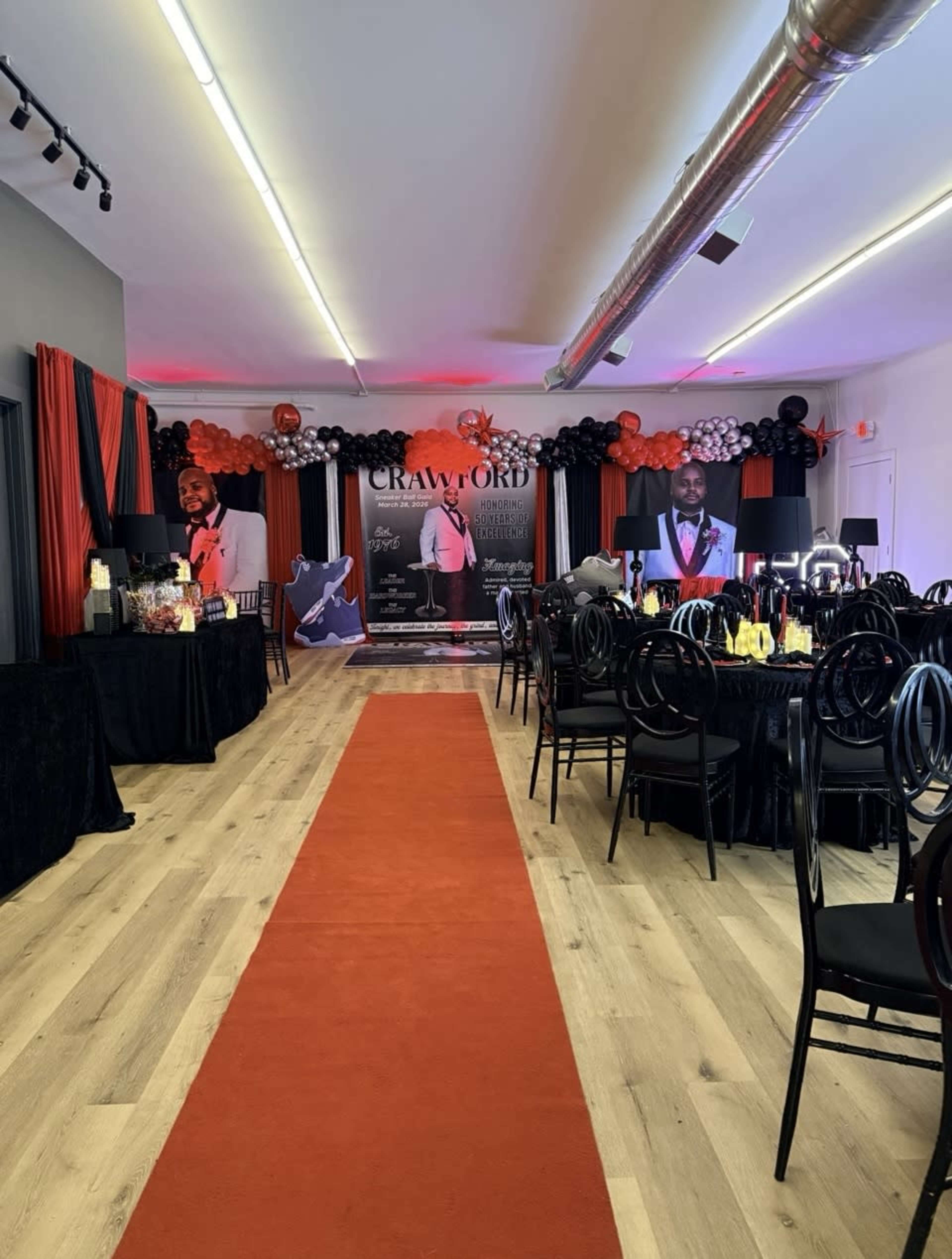 The image depicts a decorated event hall featuring a red carpet leading to a backdrop with black and red balloons, tables set with bottles and glasses, and promotional displays of a person named Crawford.