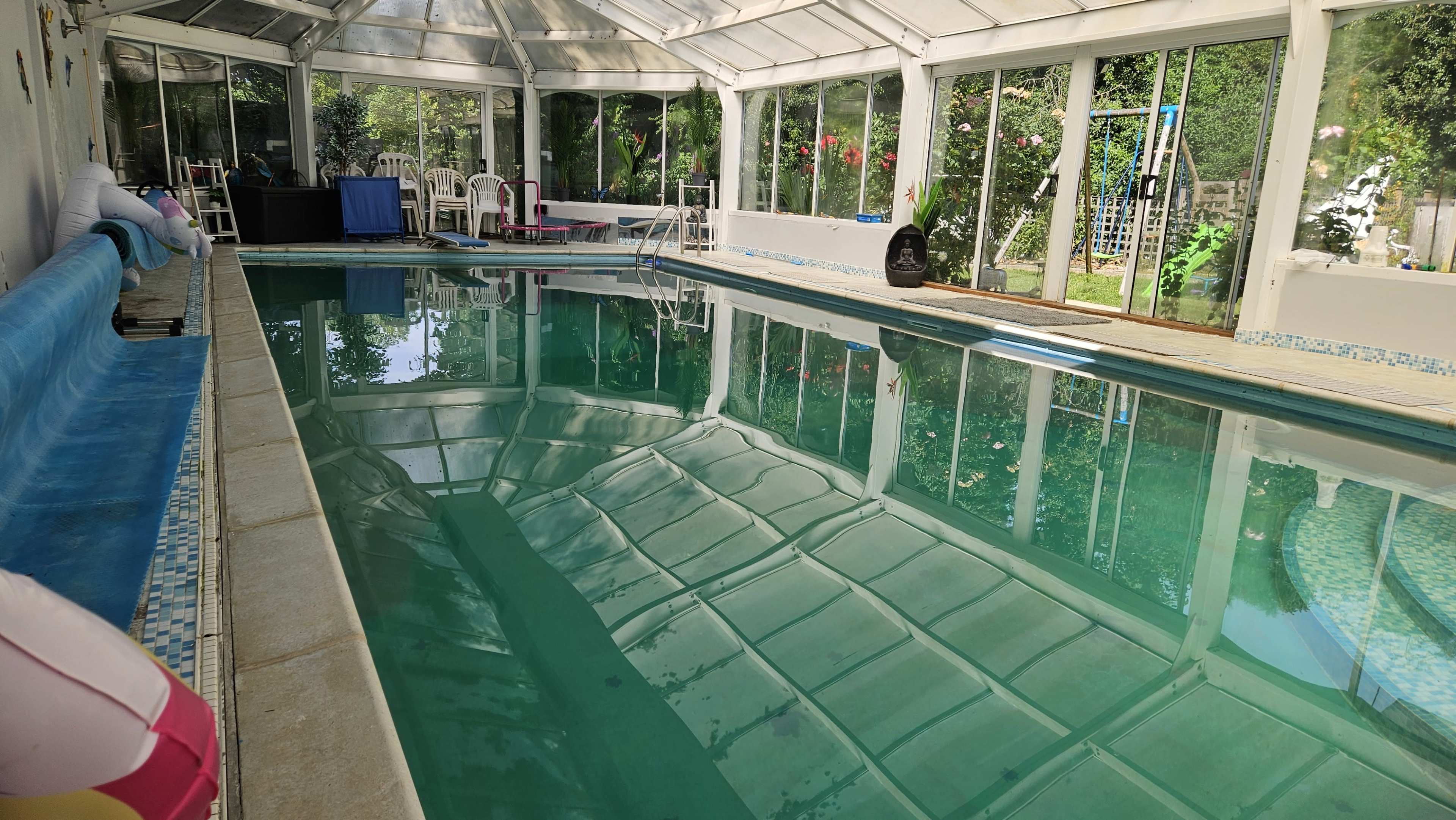The image shows an indoor swimming pool enclosed by glass walls and a ceiling, surrounded by chairs and greenery.