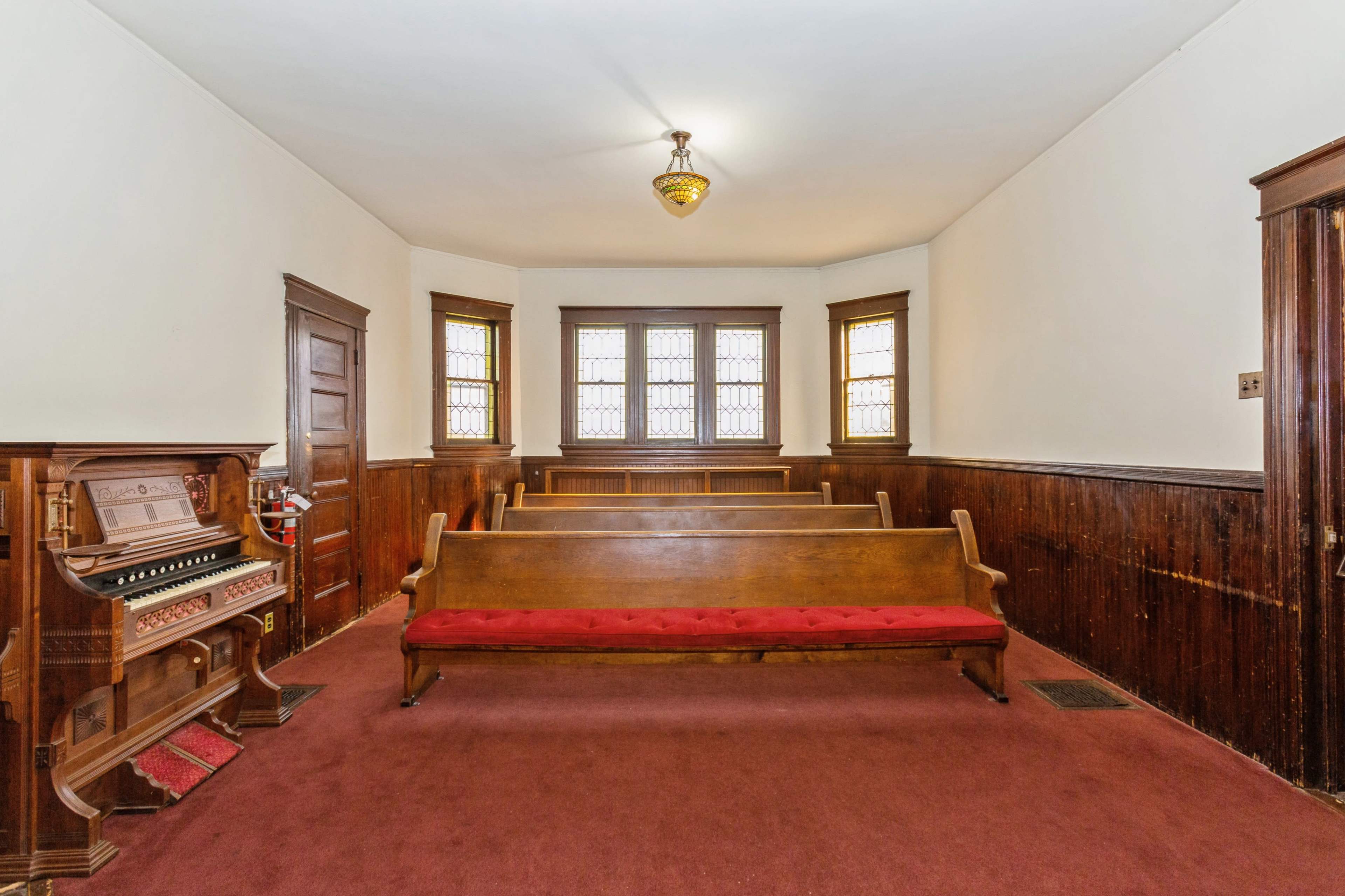 A sparsely furnished interior of a room with wooden benches, stained glass windows, and an organ in the corner.