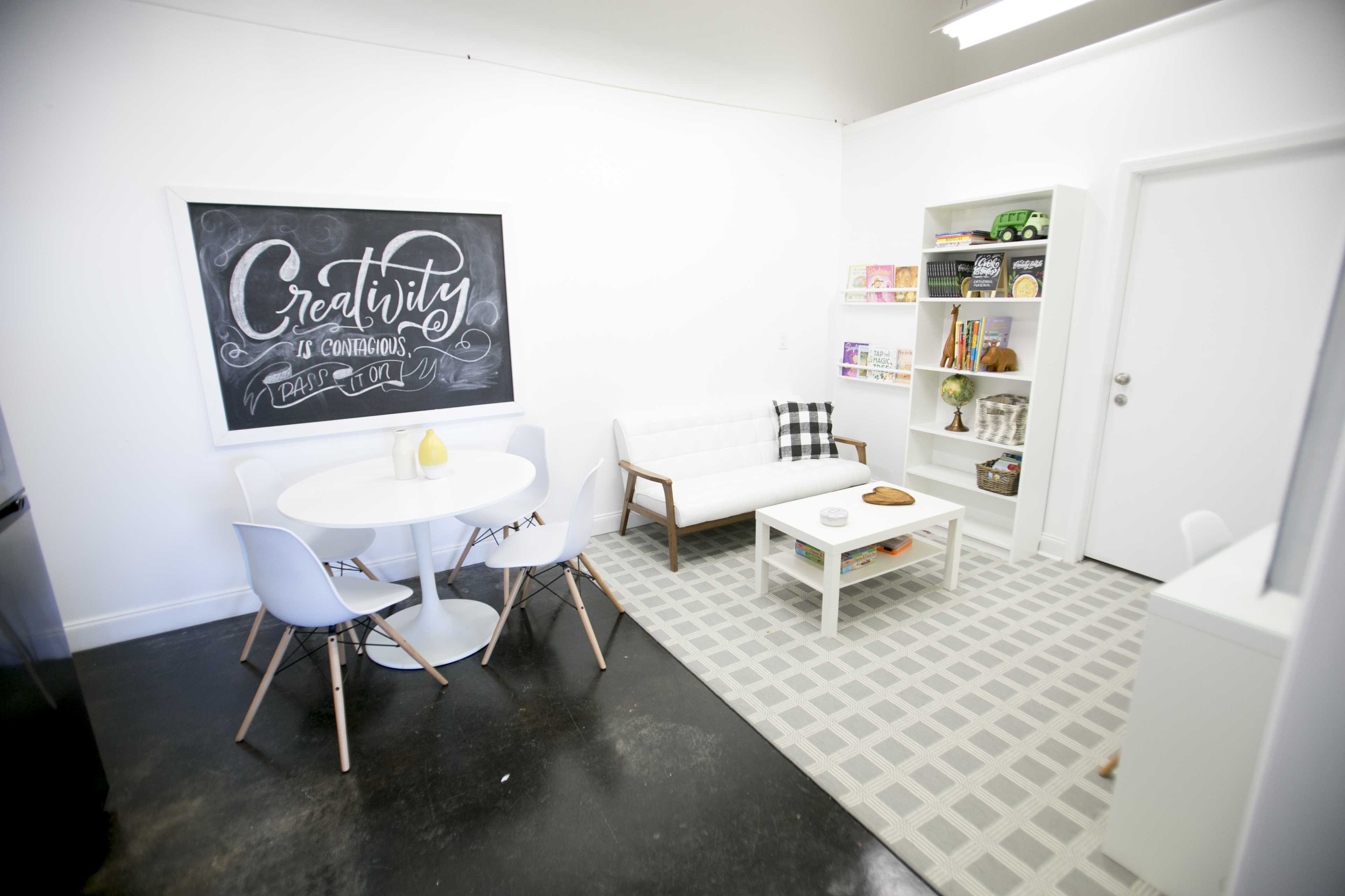 A modern, minimalist room with a round dining table, a white sofa, and a wall-mounted chalkboard featuring the word "Creativity."