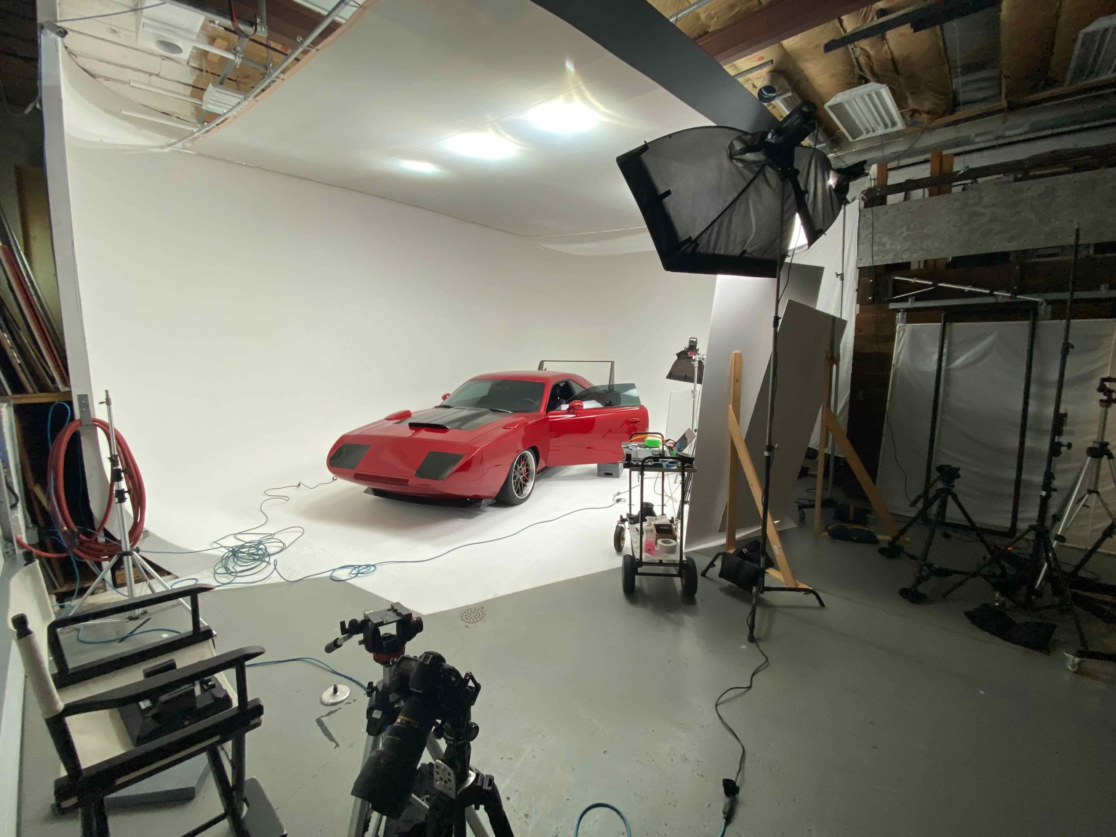A red sports car is positioned on a white backdrop in a photography studio, surrounded by various lighting equipment and tripods.