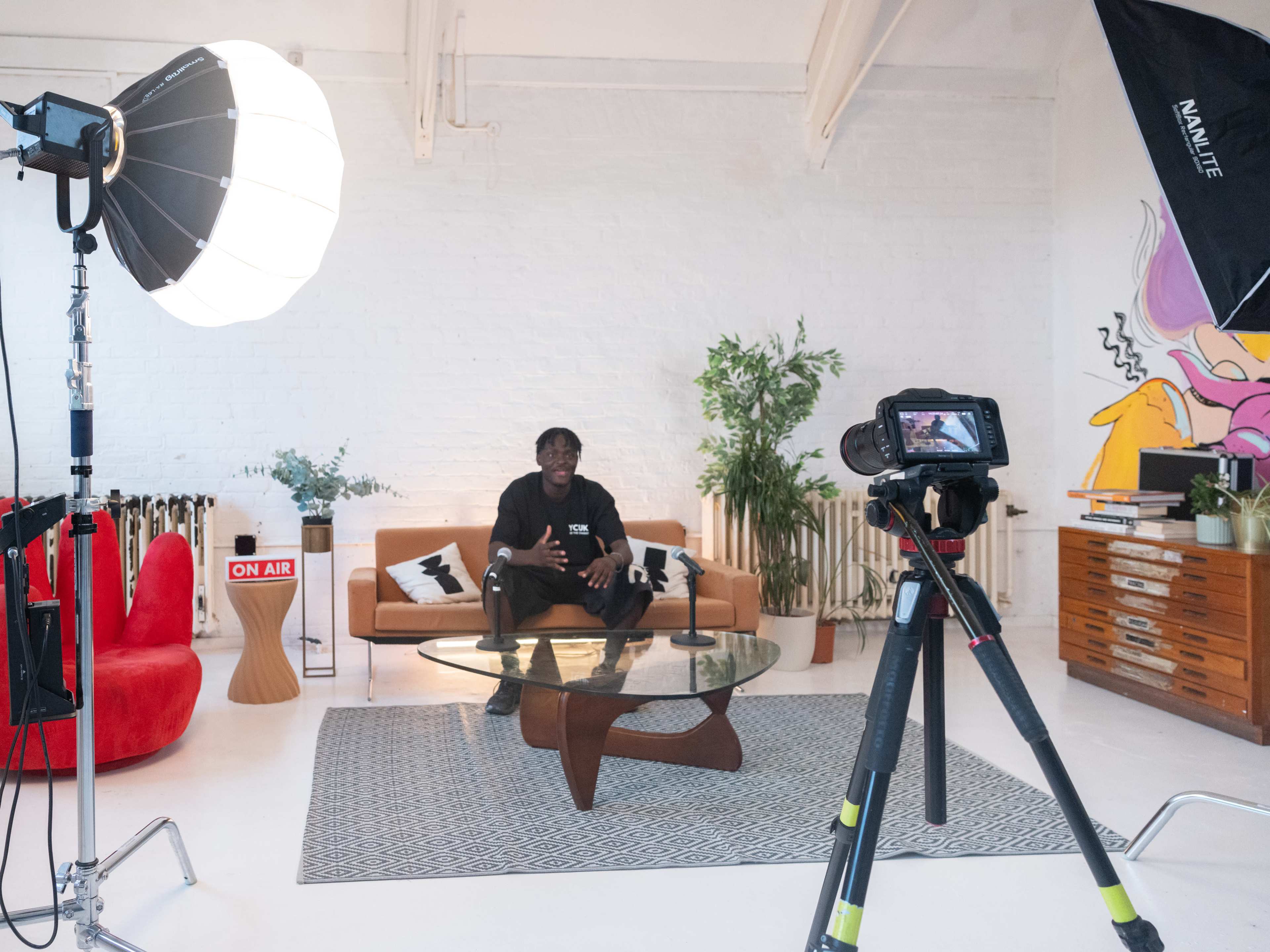 A person sits on a sofa in a well-lit studio setting with cameras, lighting equipment, and decorative plants surrounding them.