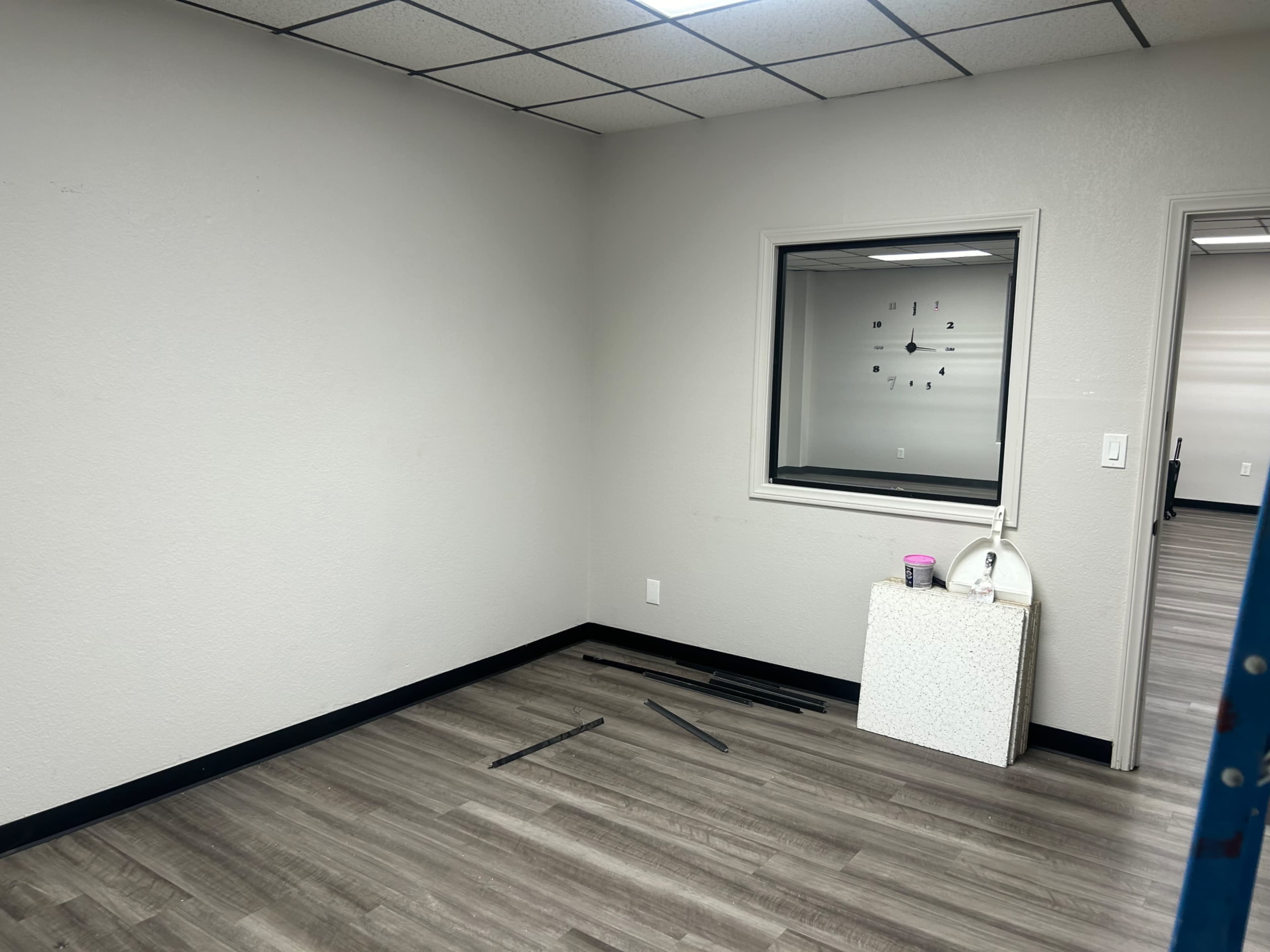 The image shows an empty office room with a clock on the wall, a stack of metal rods on the floor, and a white decorative object on a small table.