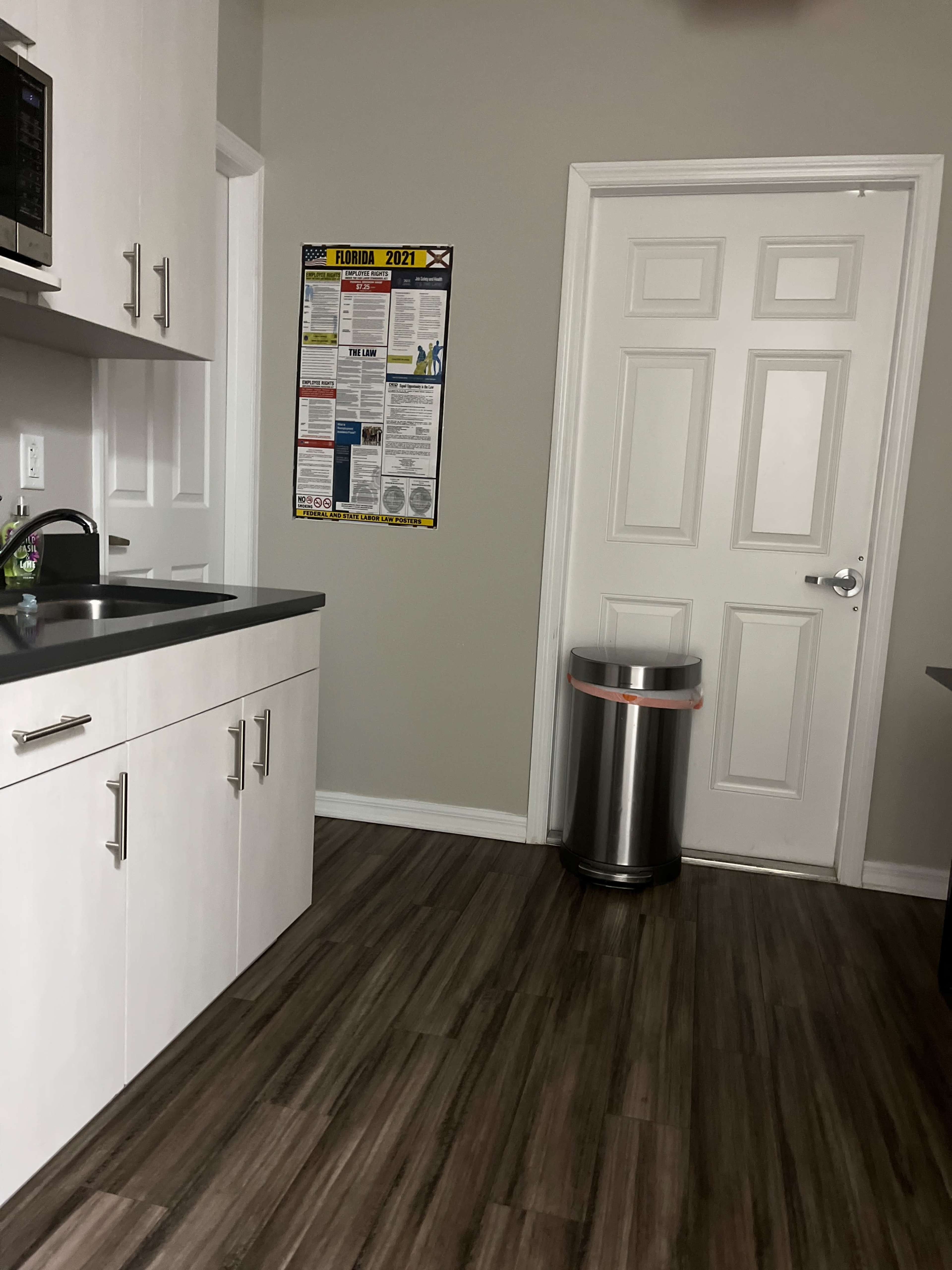A kitchen corner features a sink and countertop, a poster on the wall, and a trash can beside a closed door.