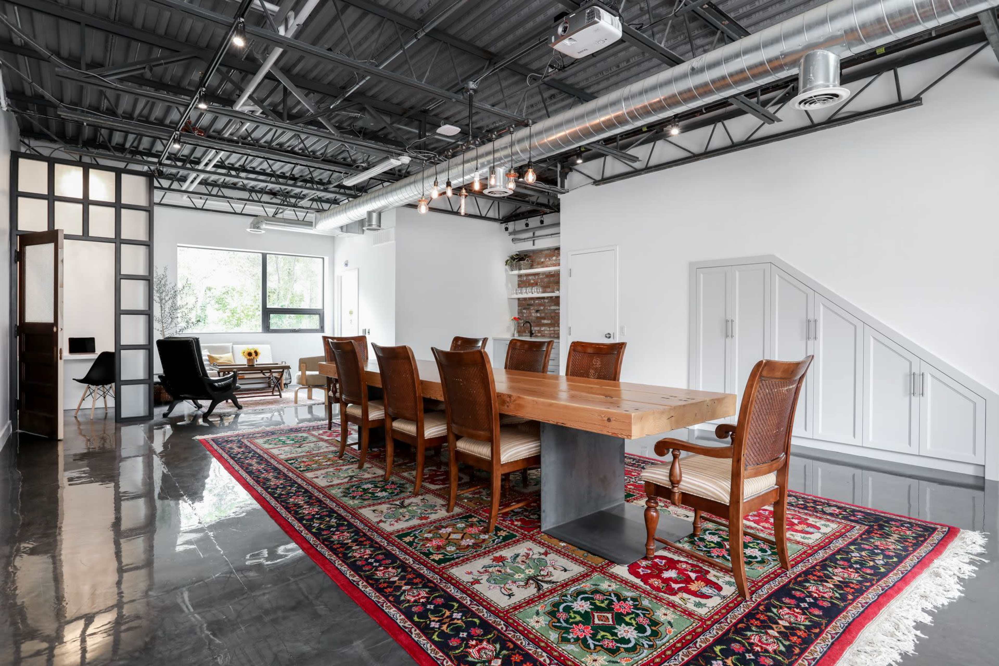 A spacious dining area features a large wooden table surrounded by eight wooden chairs on a patterned rug, with large windows and industrial-style fixtures.