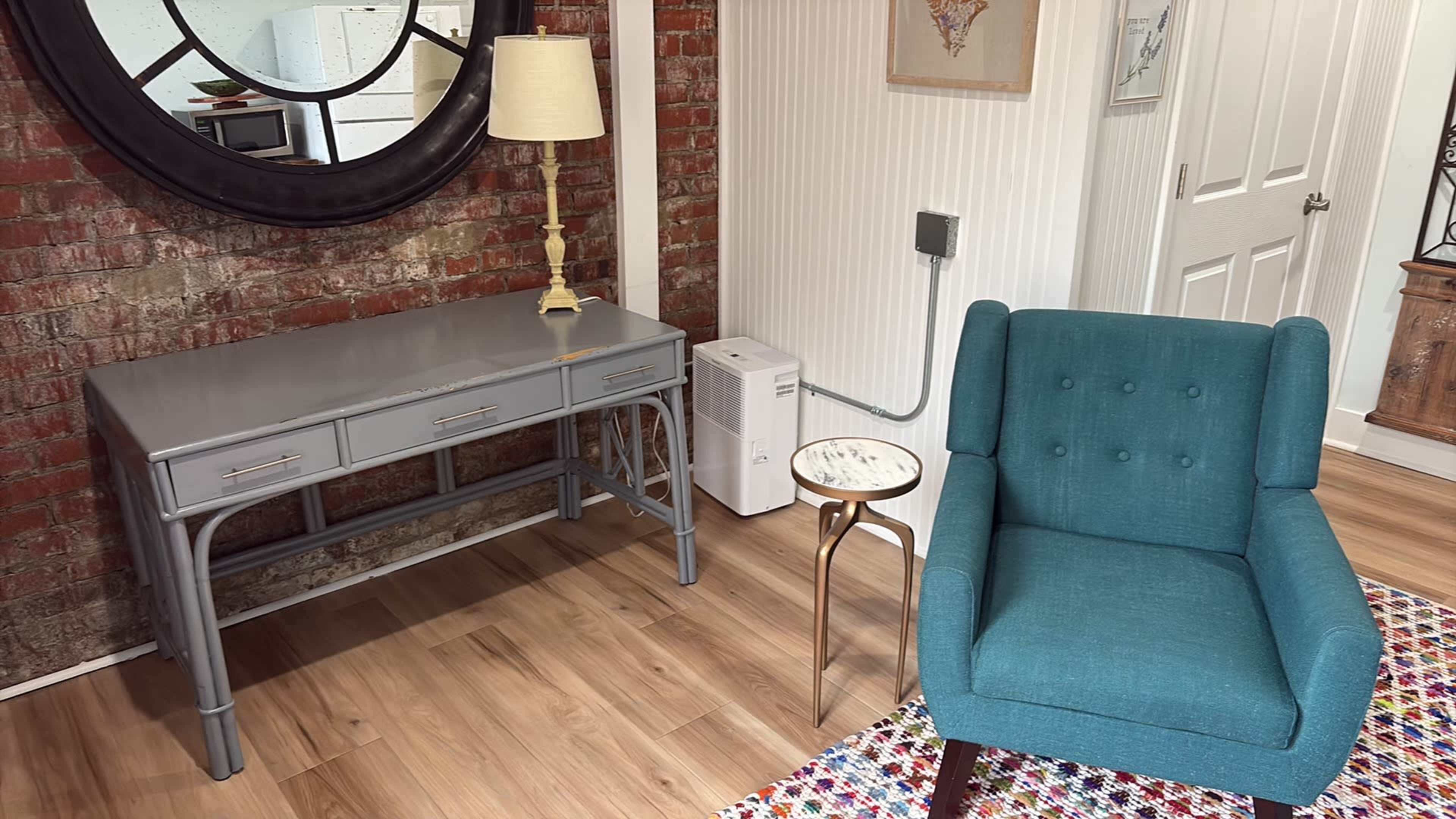 The image shows a stylish interior space featuring a gray console table with three drawers, a blue armchair, a decorative side table, and a brick wall backdrop.