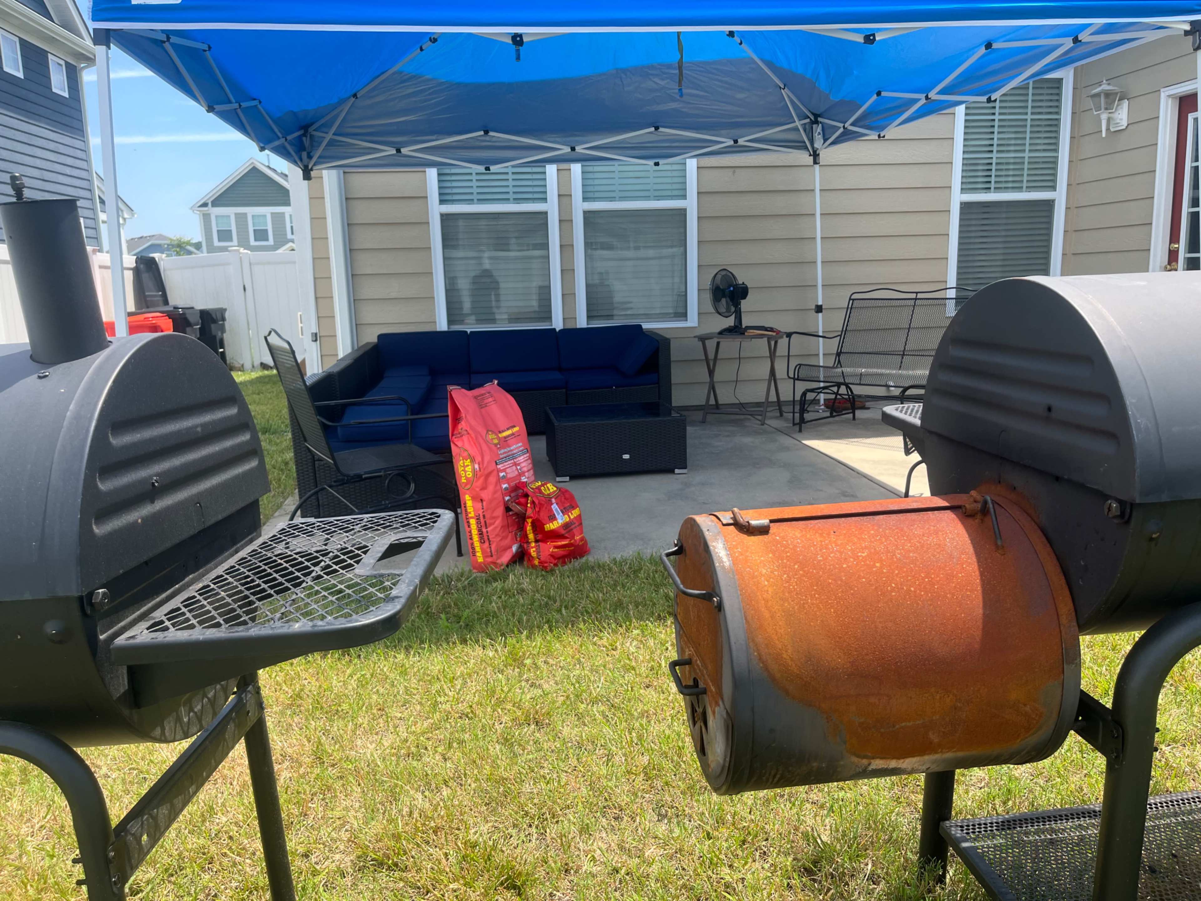 Two barbecue grills positioned in a grassy yard beneath a blue canopy, with a patio area featuring a seating arrangement and bags of charcoal nearby.