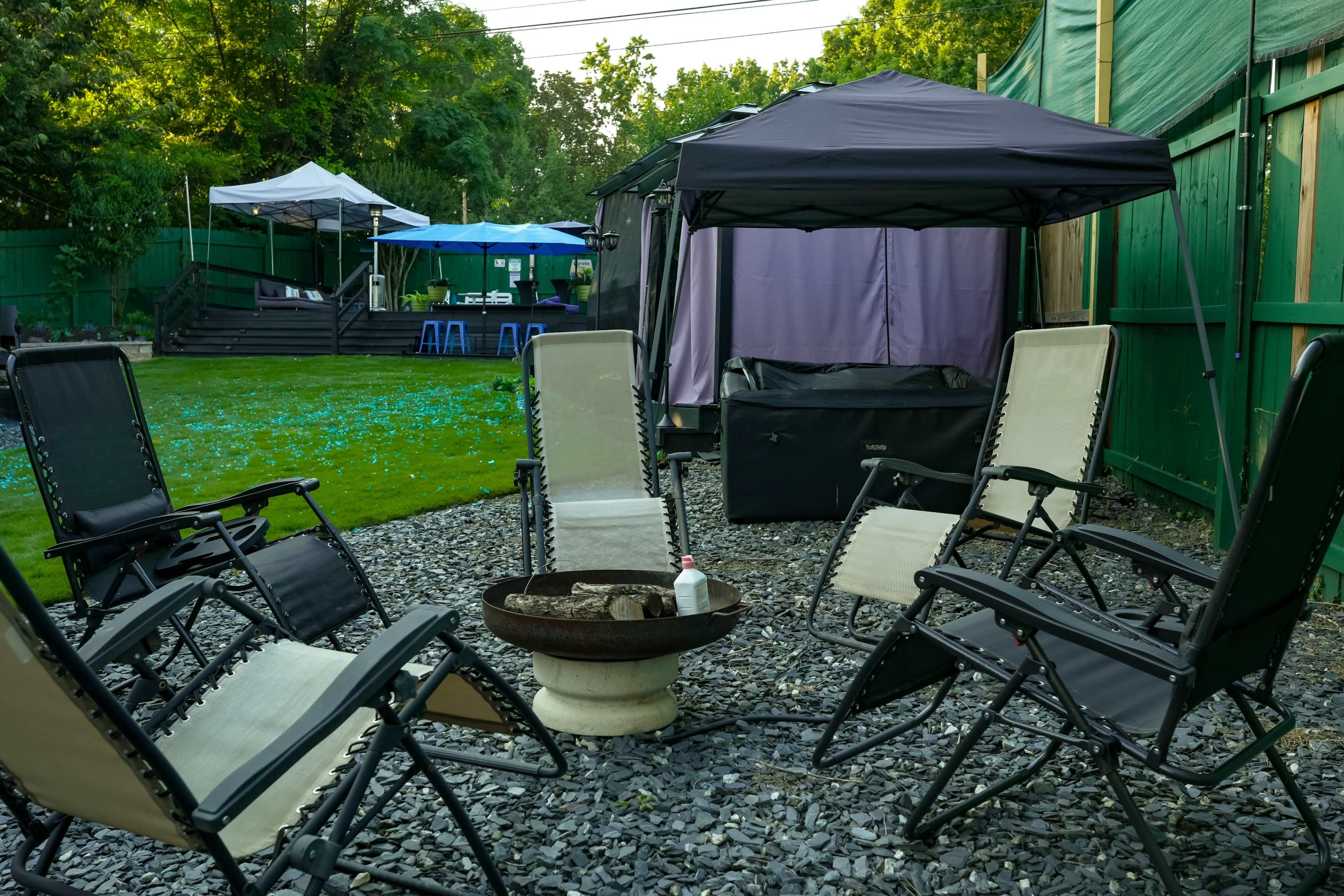 The image shows a backyard gathering area with four chairs arranged around a fire pit, surrounded by a lawn and various canopies.