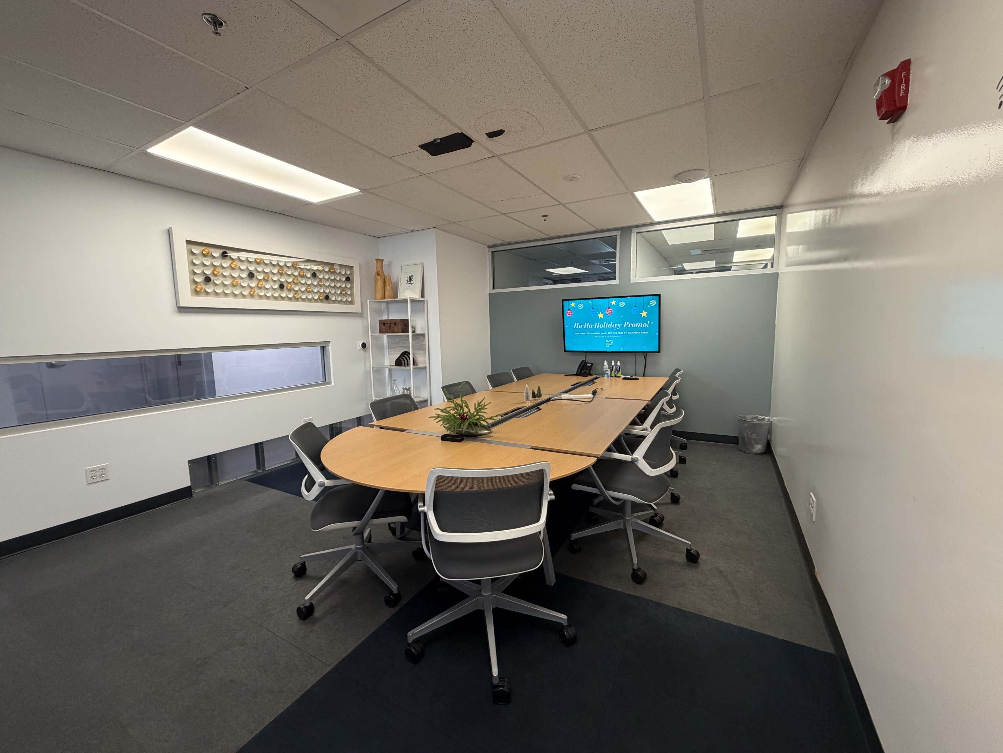 The image shows a modern conference room with a long table surrounded by ergonomic chairs and a large screen displaying a presentation on the wall.