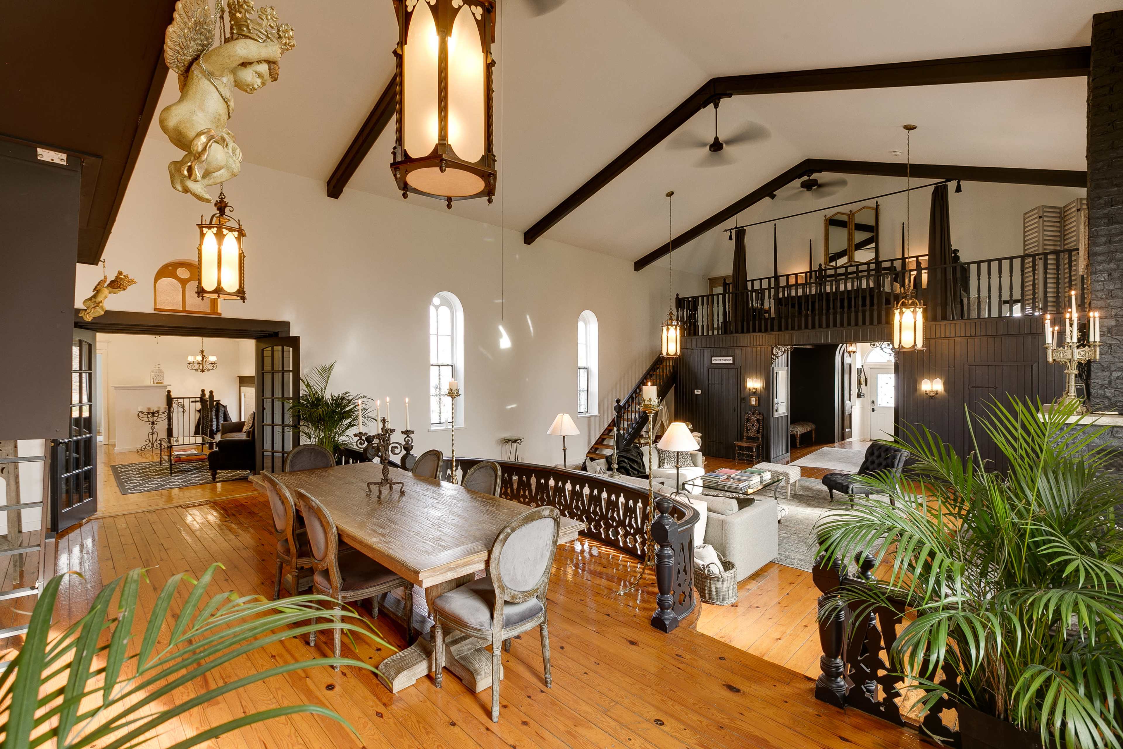The image shows a large, open-plan living area with a dining table, decorative lighting fixtures, and a staircase leading to a lofted space.