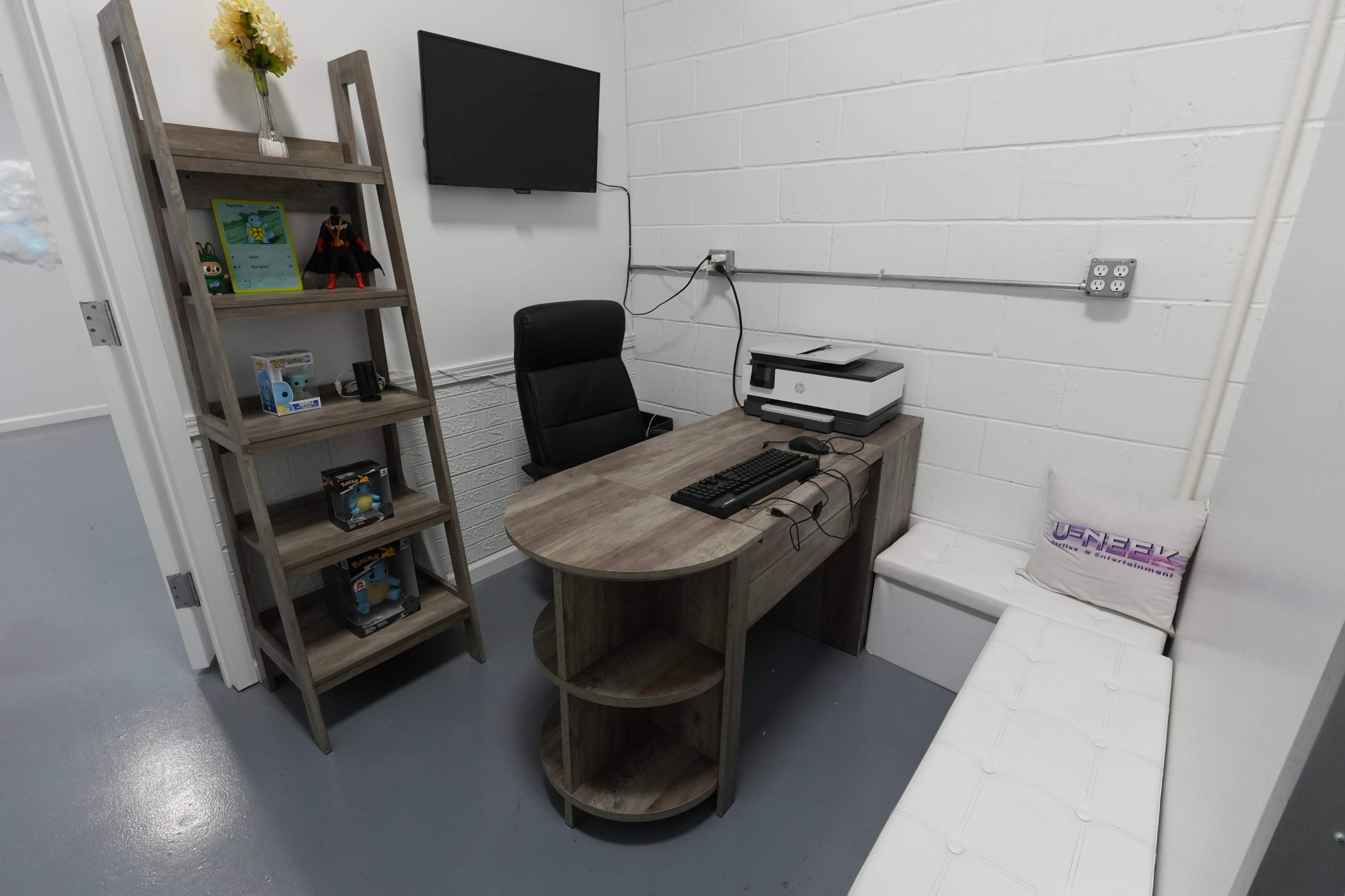The image shows a small office space with a desk, an ergonomic chair, a printer, and a shelving unit displaying various items, along with a wall-mounted television.