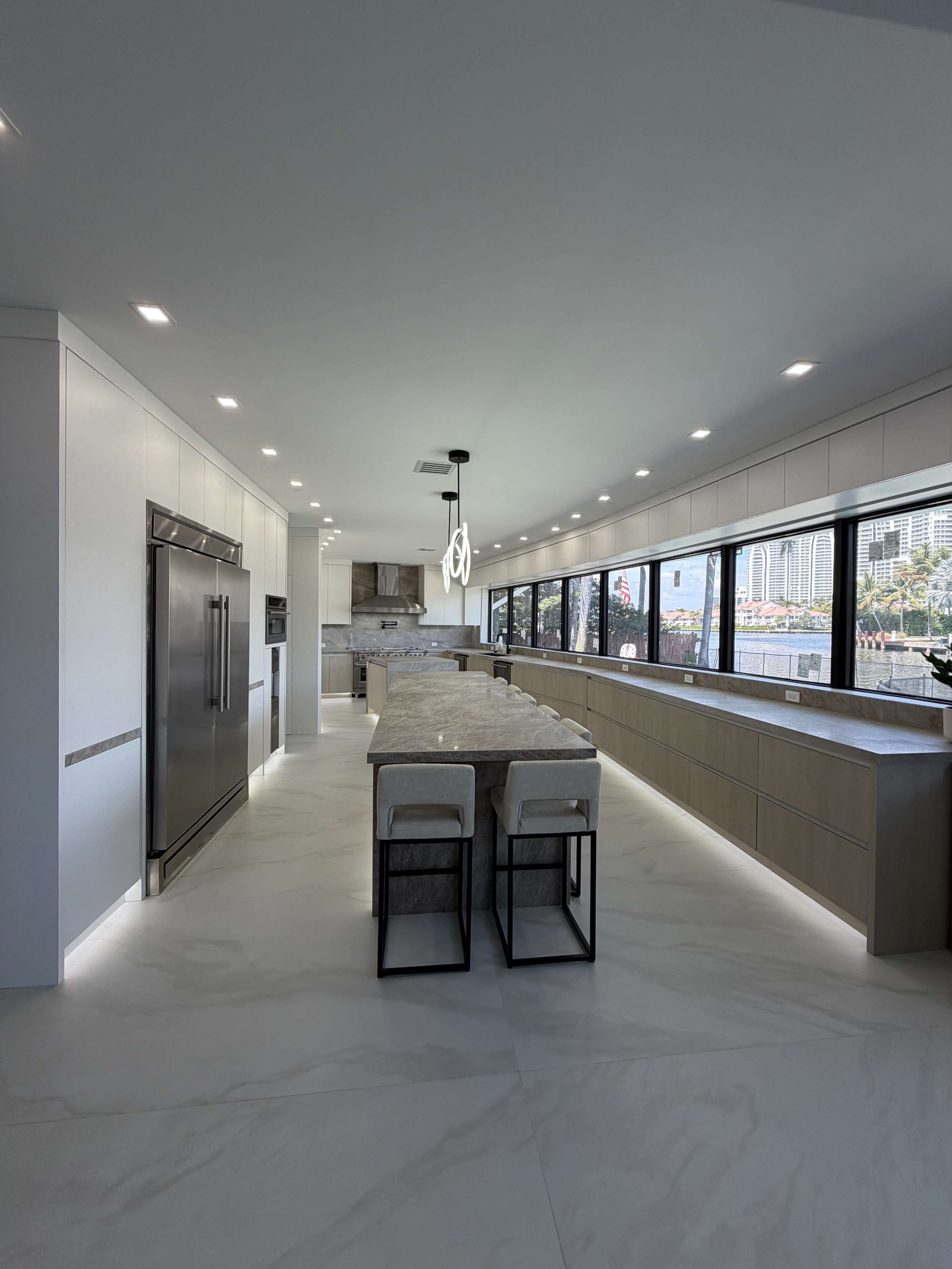 A modern kitchen features sleek cabinetry, a large marble island with bar stools, and expansive windows that allow natural light to fill the space.