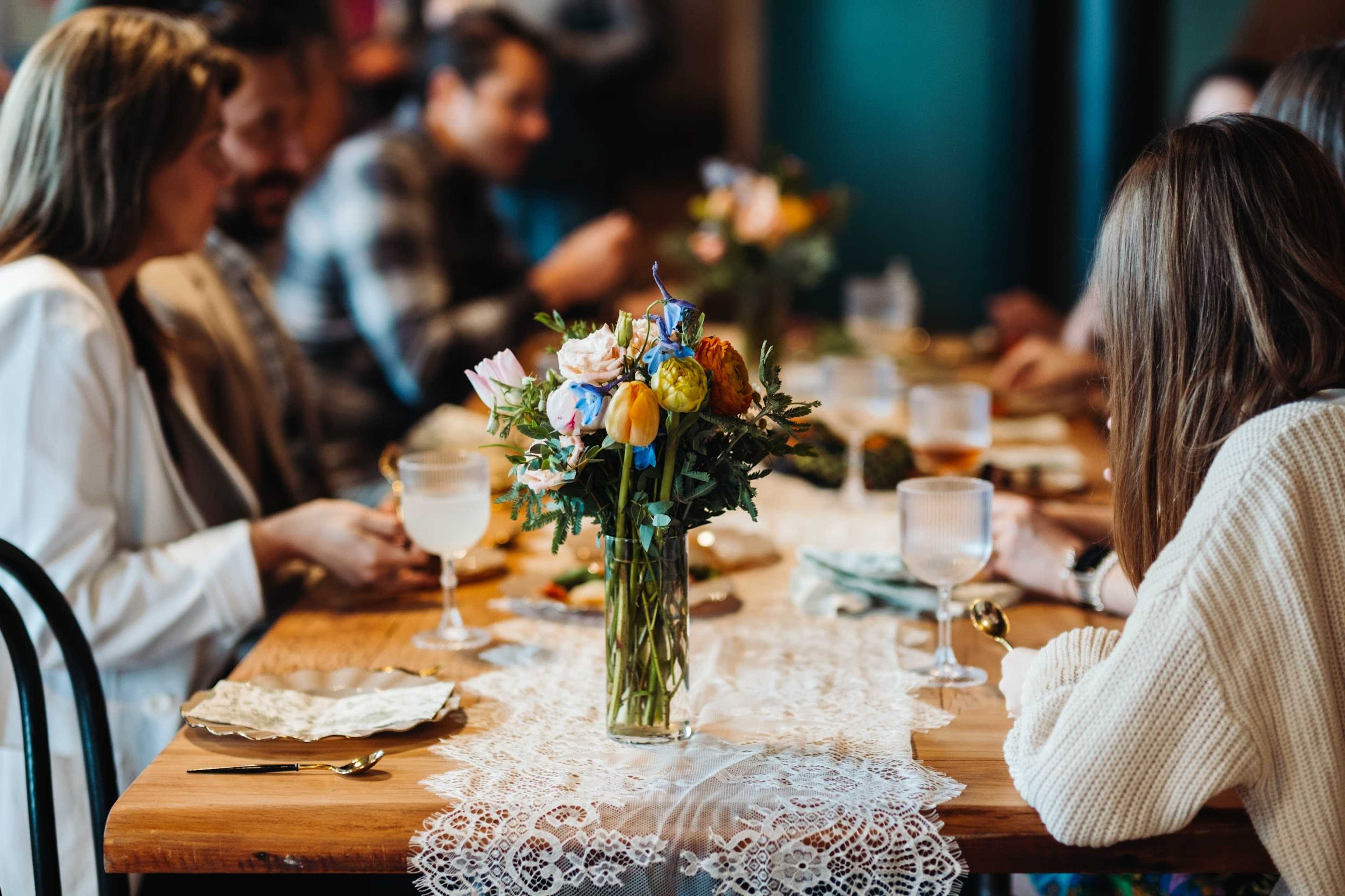 A group of people is seated around a wooden table adorned with a lace runner and a vase of flowers, enjoying a meal together.