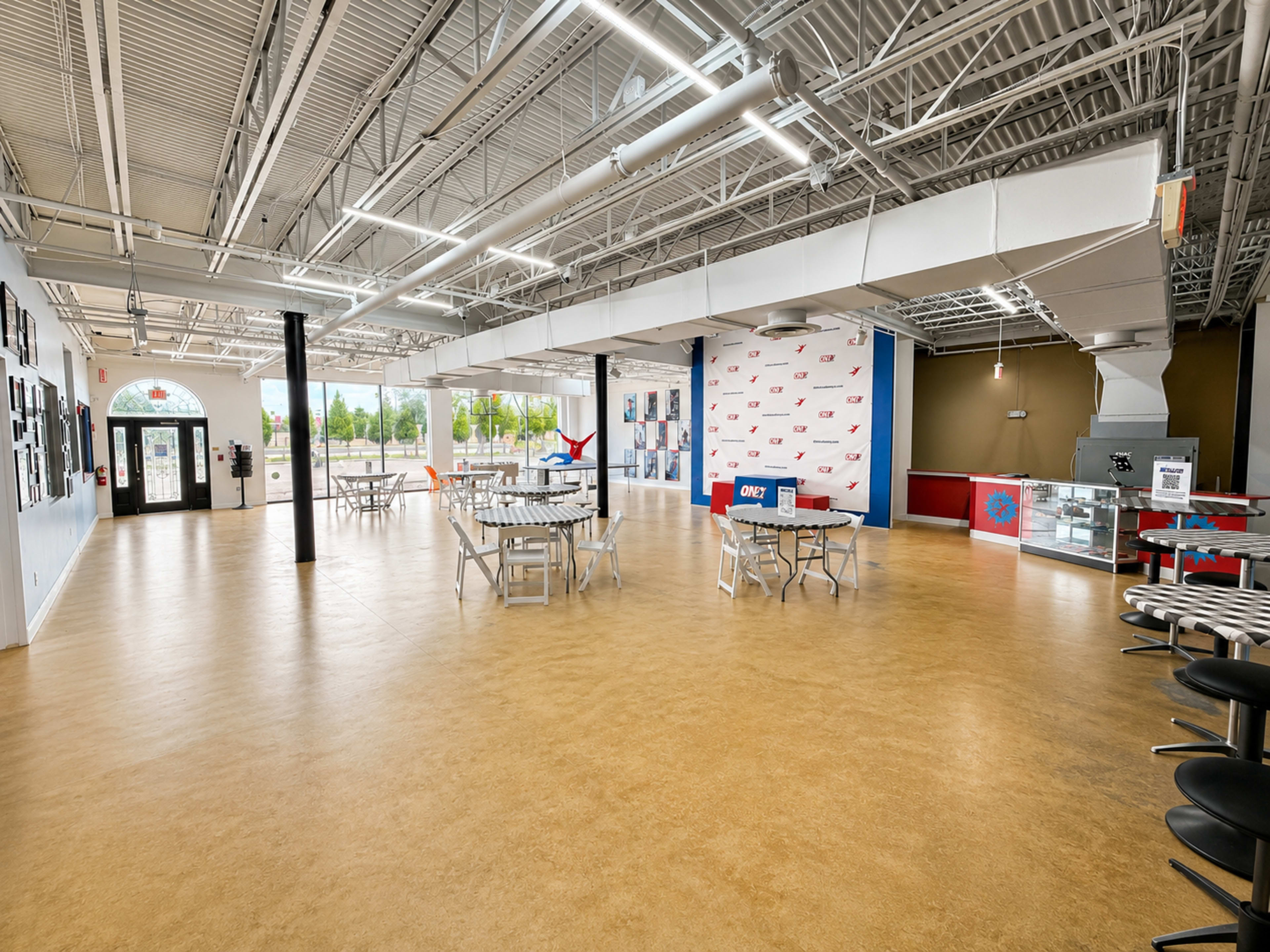The image shows an empty indoor space with tables and chairs arranged for seating, large windows allowing natural light, and a red and white wall featuring various posters.